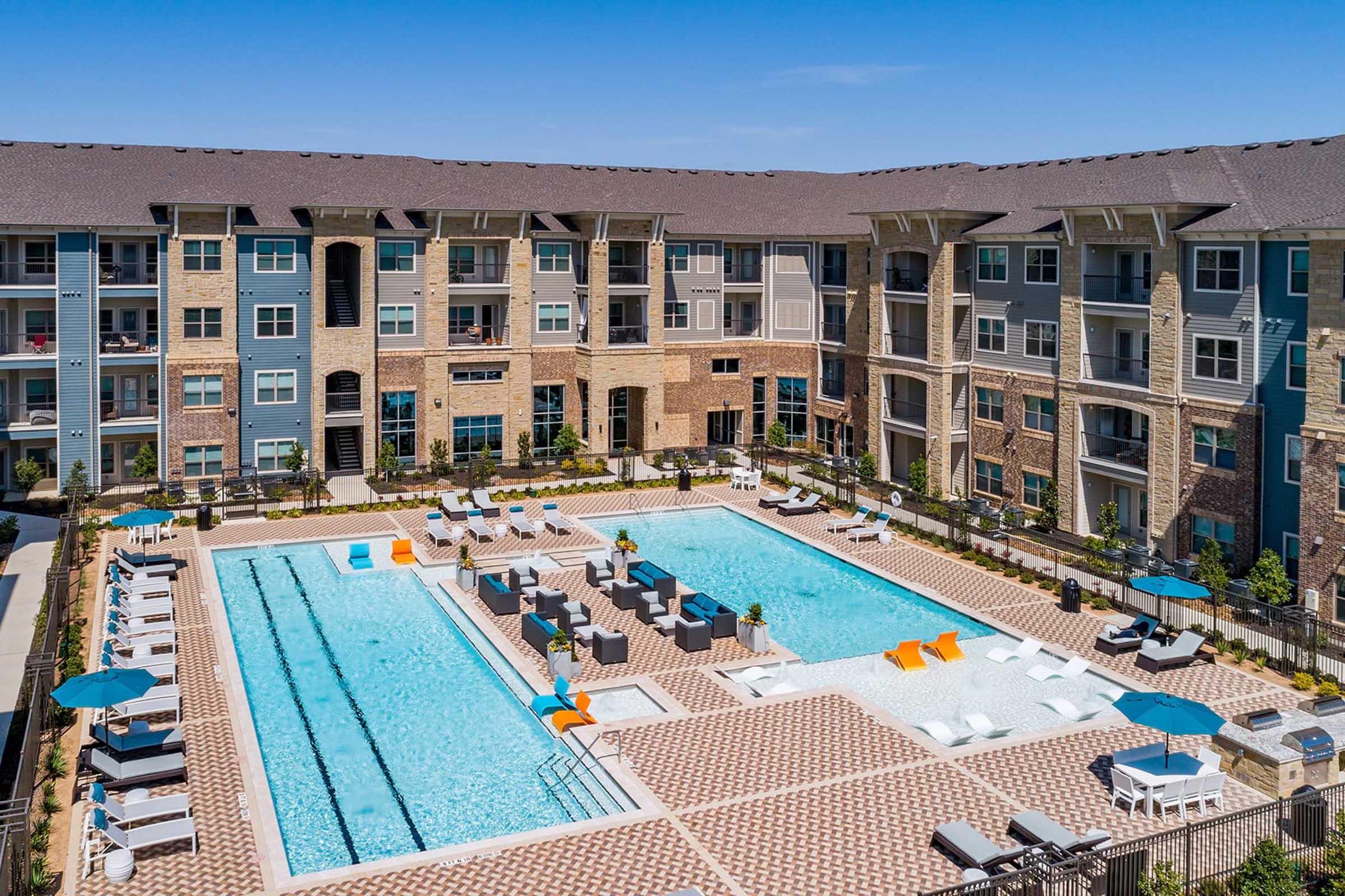 Aerial view of a modern apartment complex featuring two swimming pools surrounded by lounge chairs and umbrellas.