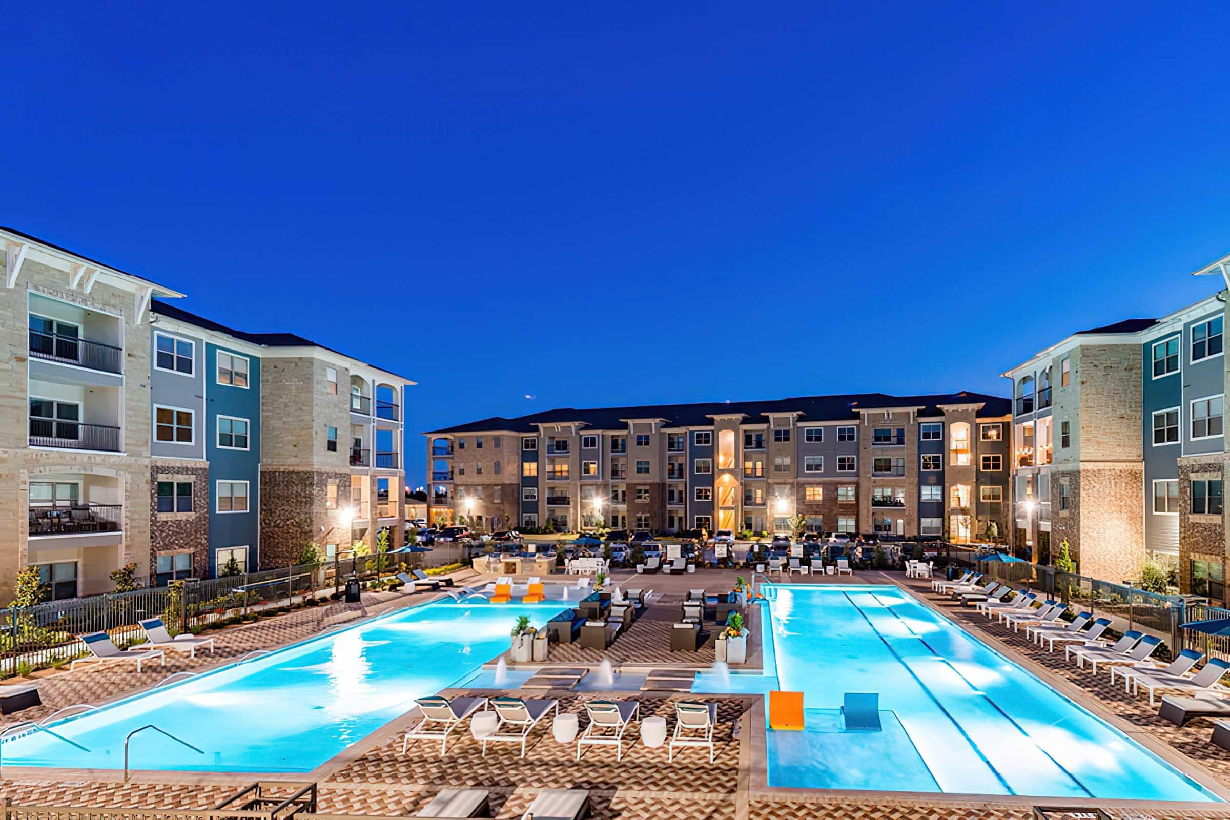 A well-lit outdoor swimming pool area at dusk, surrounded by modern multi-story apartment buildings. The scene features two large pools with lounge chairs and colorful poolside furniture, creating a relaxing ambiance with a clear blue sky in the background.