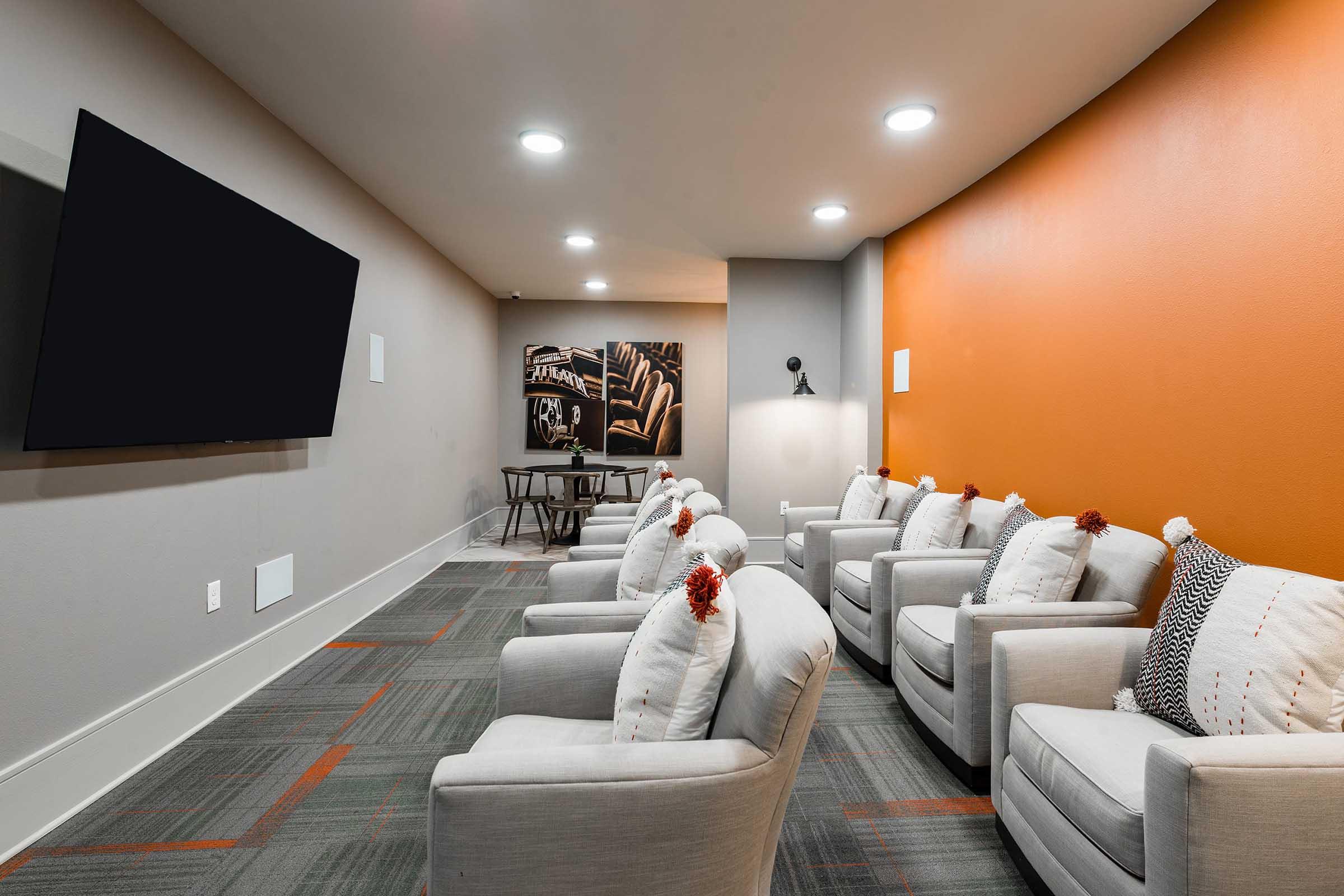 Cozy seating area with several light gray armchairs decorated with pillows, a large flat-screen TV mounted on the wall, and an orange accent wall.