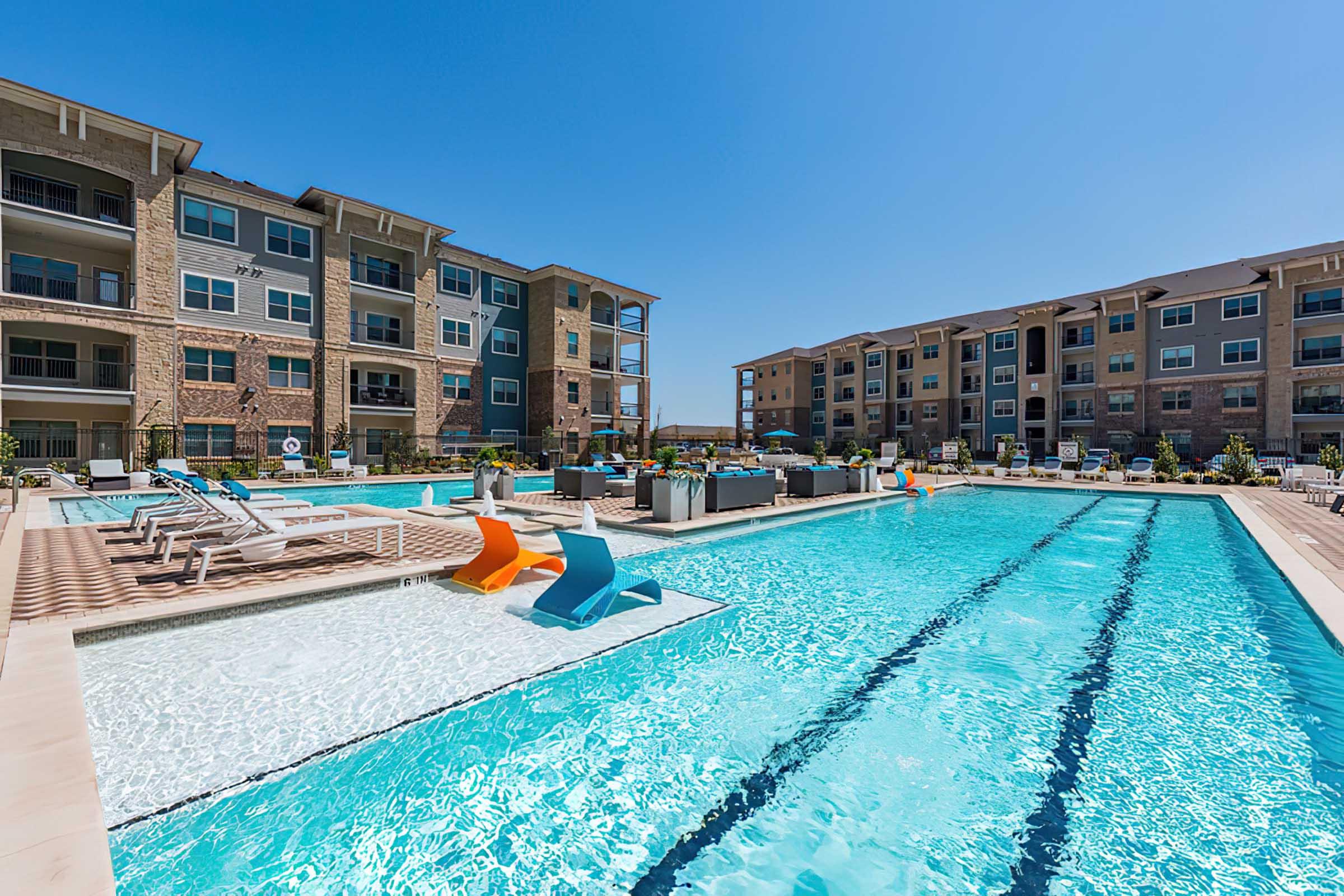A large outdoor swimming pool surrounded by lounge chairs and colorful poolside seating. Modern apartment buildings are visible in the background under a clear blue sky.