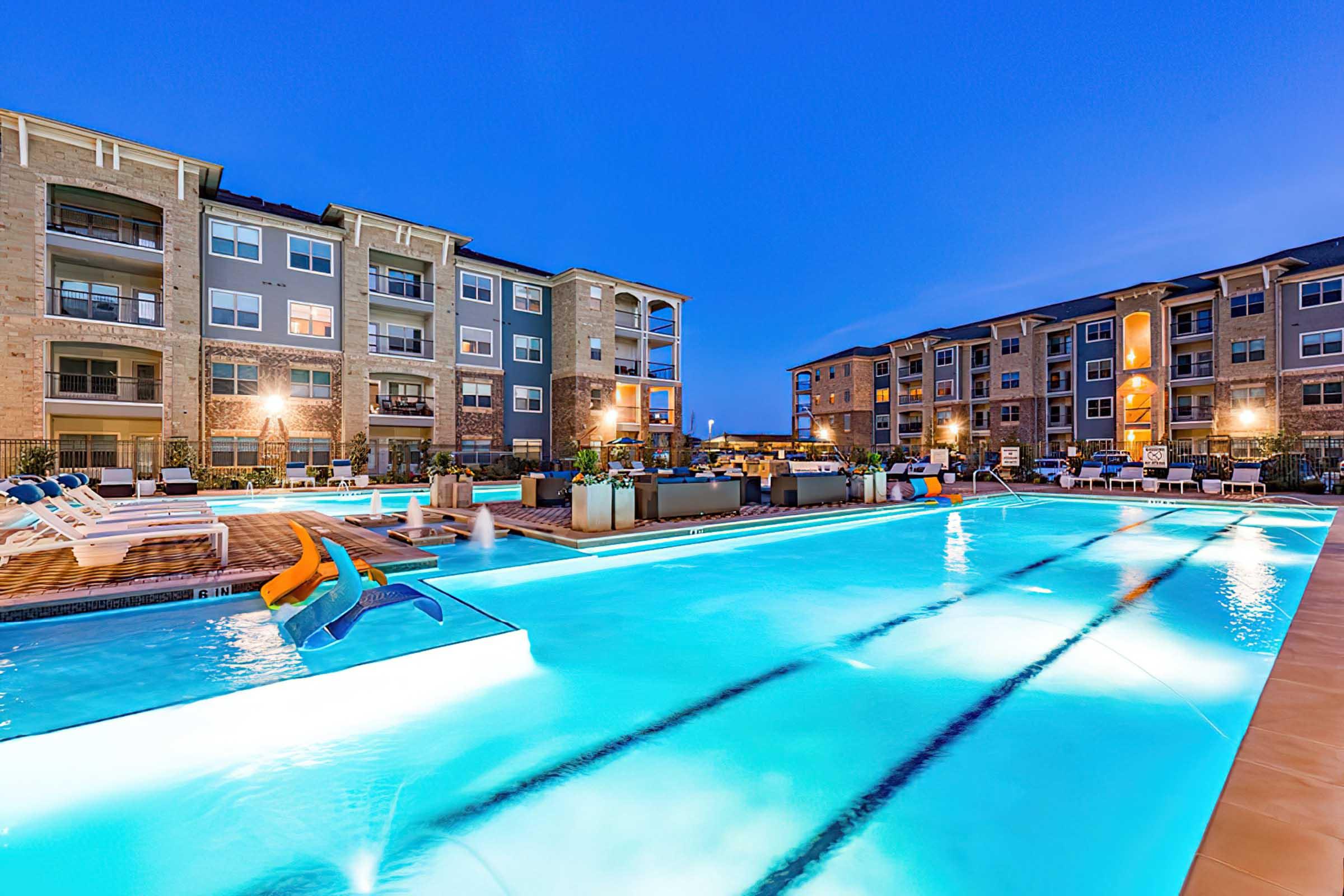A clear blue swimming pool surrounded by modern apartment buildings at dusk. Lounge chairs line the poolside, and soft lighting illuminates the area, creating a relaxing atmosphere.