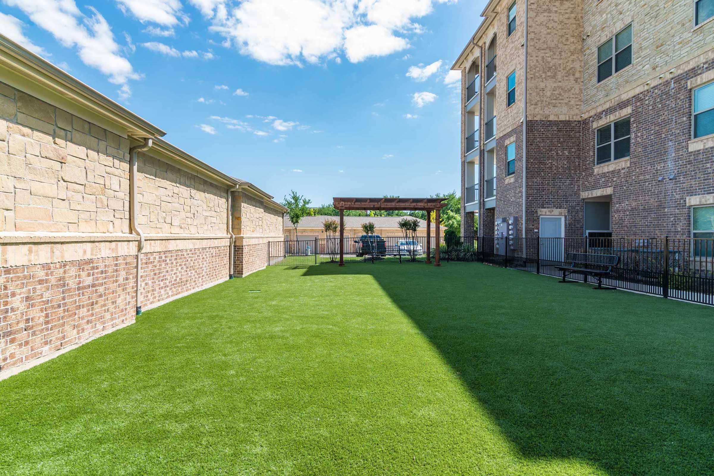 A green lawn area surrounded by buildings, featuring a pergola against a clear blue sky.
