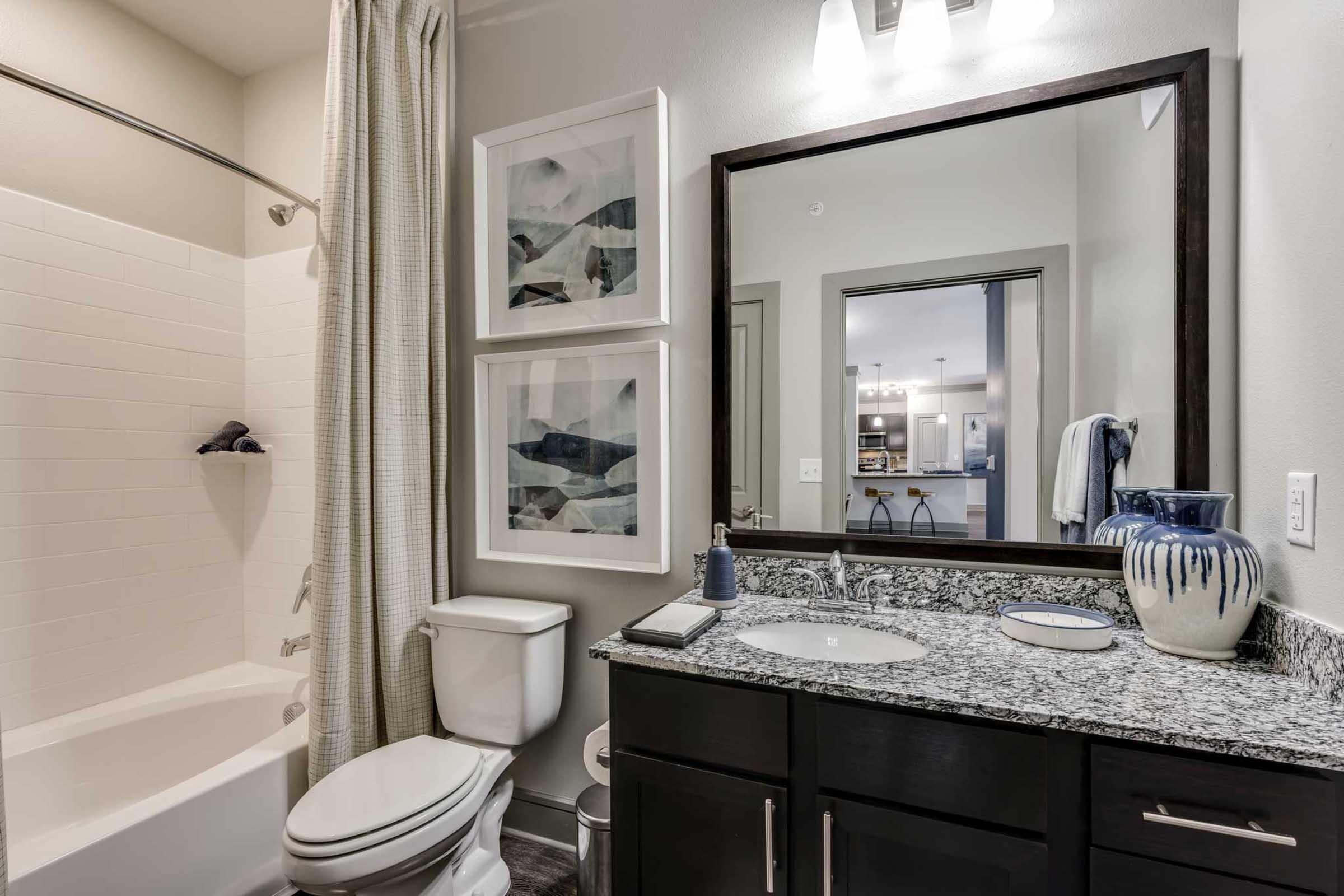 A modern bathroom featuring a white bathtub with a shower, a dark wood vanity with a granite countertop, and a mirror.