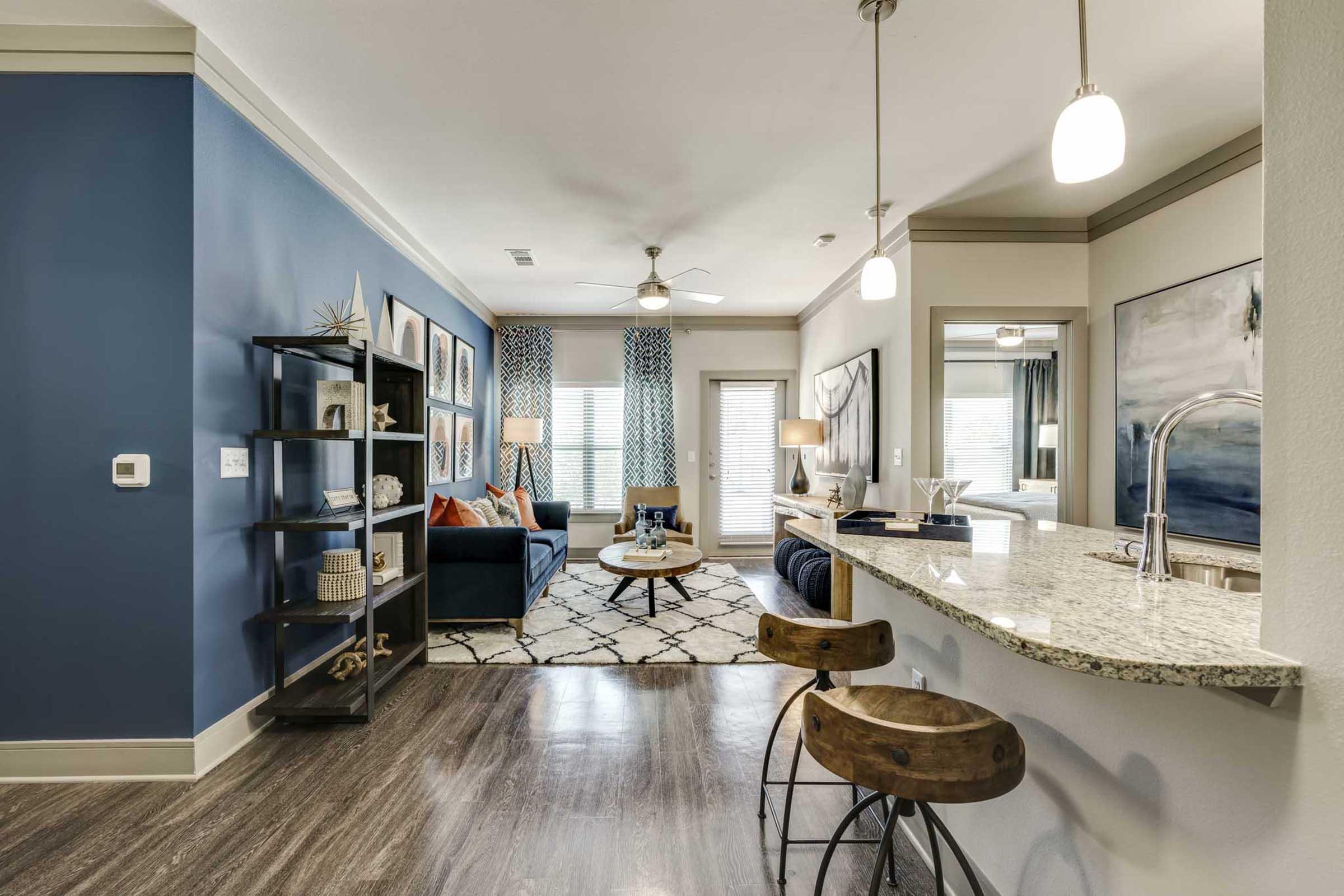 A stylish modern living room and kitchen area featuring a blue accent wall, a comfortable sofa, decorative artwork, and a dining space with bar stools.