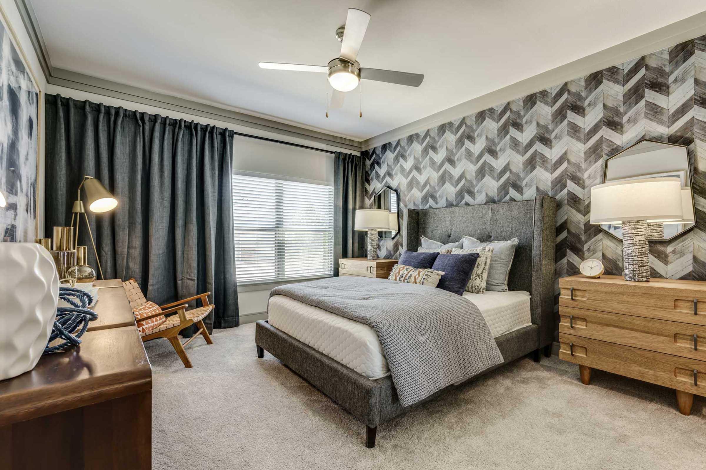 A cozy bedroom featuring a gray upholstered bed with decorative pillows, a stylish herringbone-patterned accent wall, wooden furniture, and large windows covered with dark curtains.