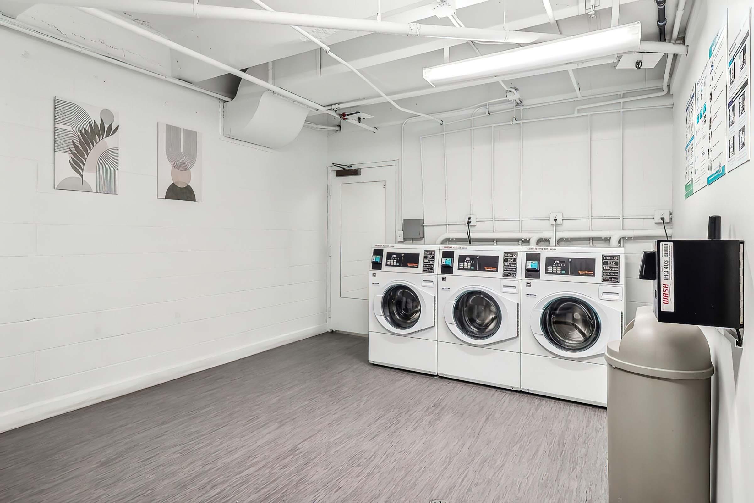 A bright and clean laundry room featuring three front-loading washing machines. The walls are painted white, with simple artwork displayed above the machines. There is a trash bin on the right, and the space is well-lit with overhead lights, creating a modern and inviting atmosphere for doing laundry.