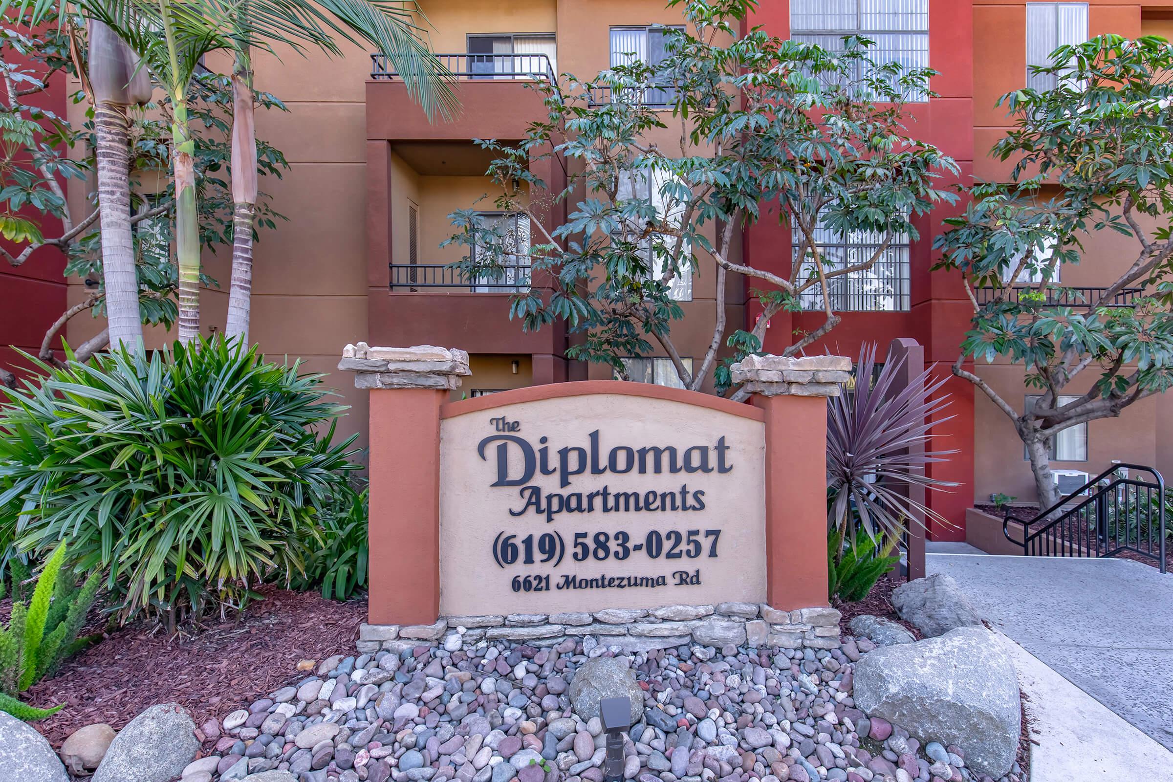 Sign for The Diplomat Apartments, featuring the name and contact information. Surrounding the sign are various plants and landscaping, with the building façade visible in the background. The location is 6621 Montezuma Rd, with a stairway leading from the sign.