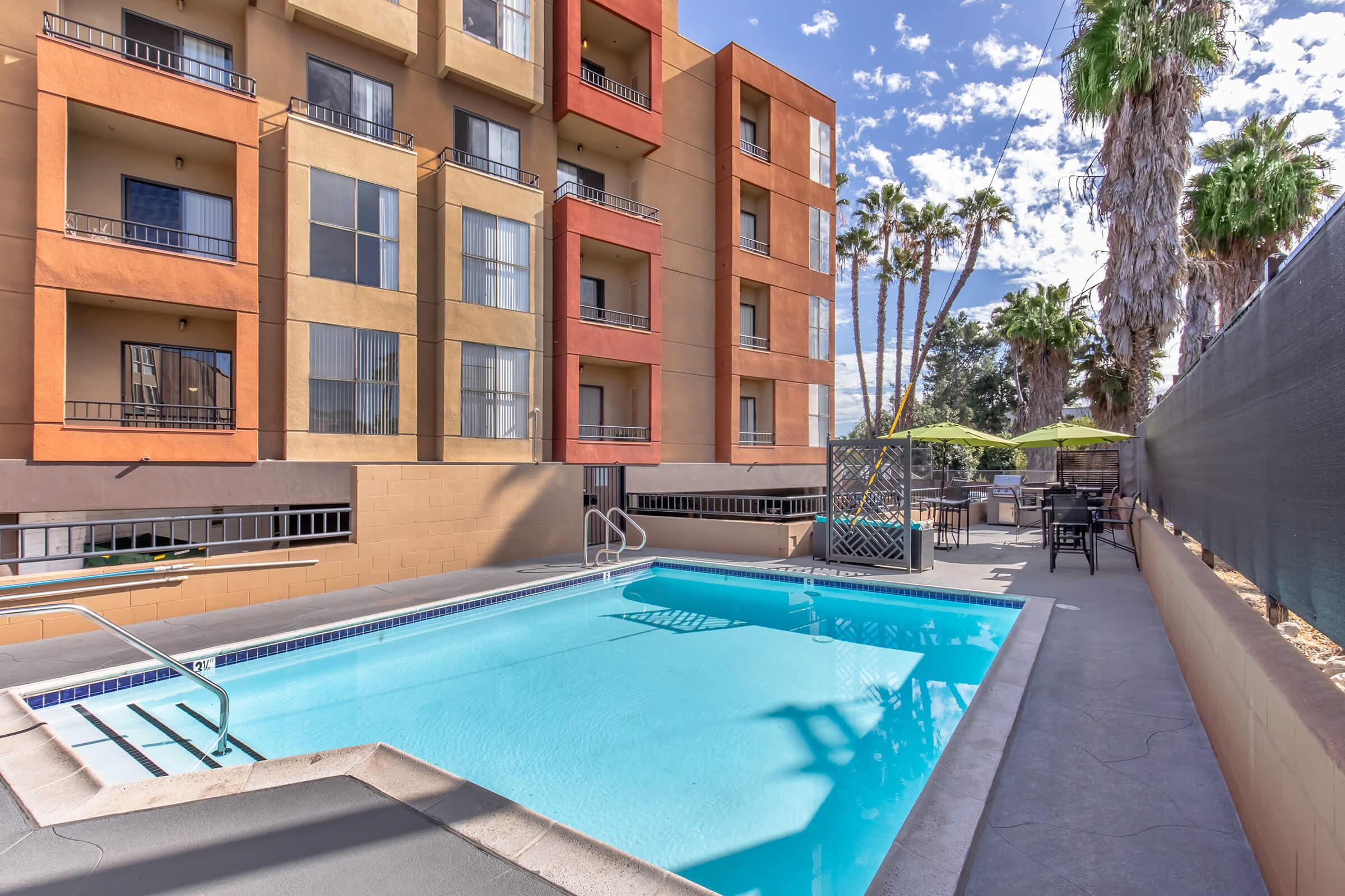 A clear blue swimming pool surrounded by a patio area with lounge chairs and green umbrellas. The pool area is bordered by a modern, multi-story building with large windows. Palm trees are visible in the background, under a partly cloudy sky.
