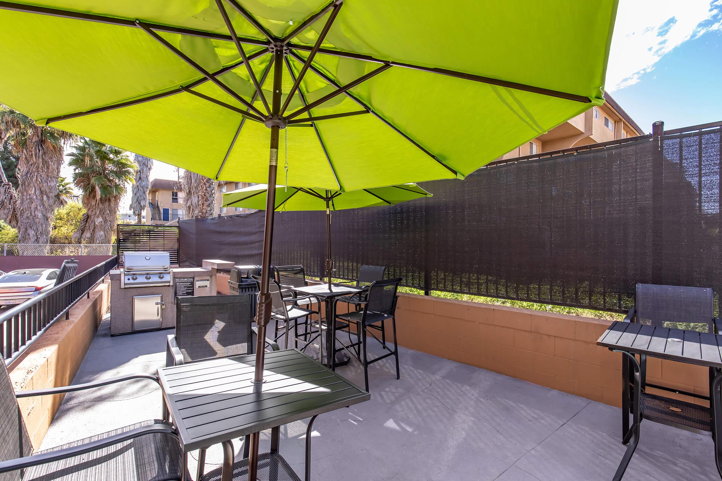 Outdoor patio area with green umbrellas providing shade over black metal tables and chairs. A built-in grill is situated against a wall, surrounded by palm trees and a privacy screen, creating a relaxing dining space. The setting is bright and inviting, ideal for gatherings.
