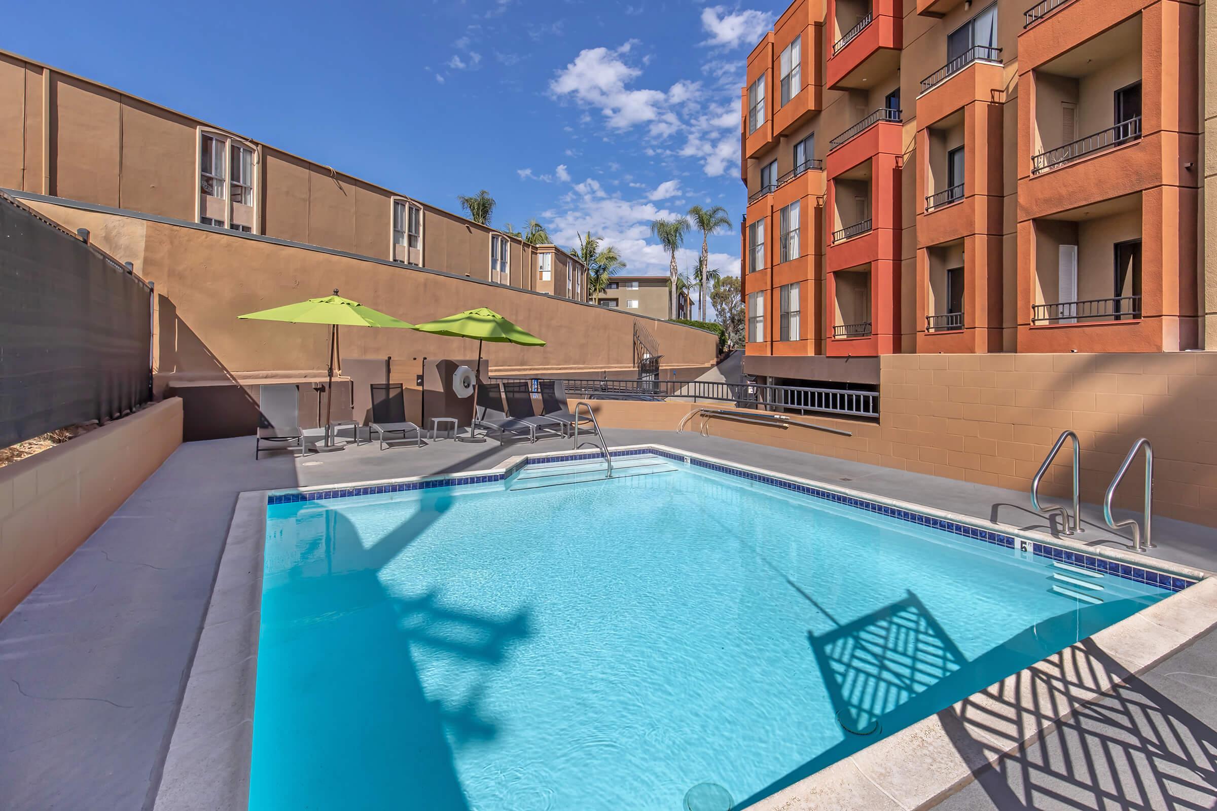 A clear blue swimming pool surrounded by lounge chairs and umbrellas in a sunny outdoor setting. Nearby, there are modern apartment buildings with balconies, and palm trees add to the tropical atmosphere under a mostly sunny sky.