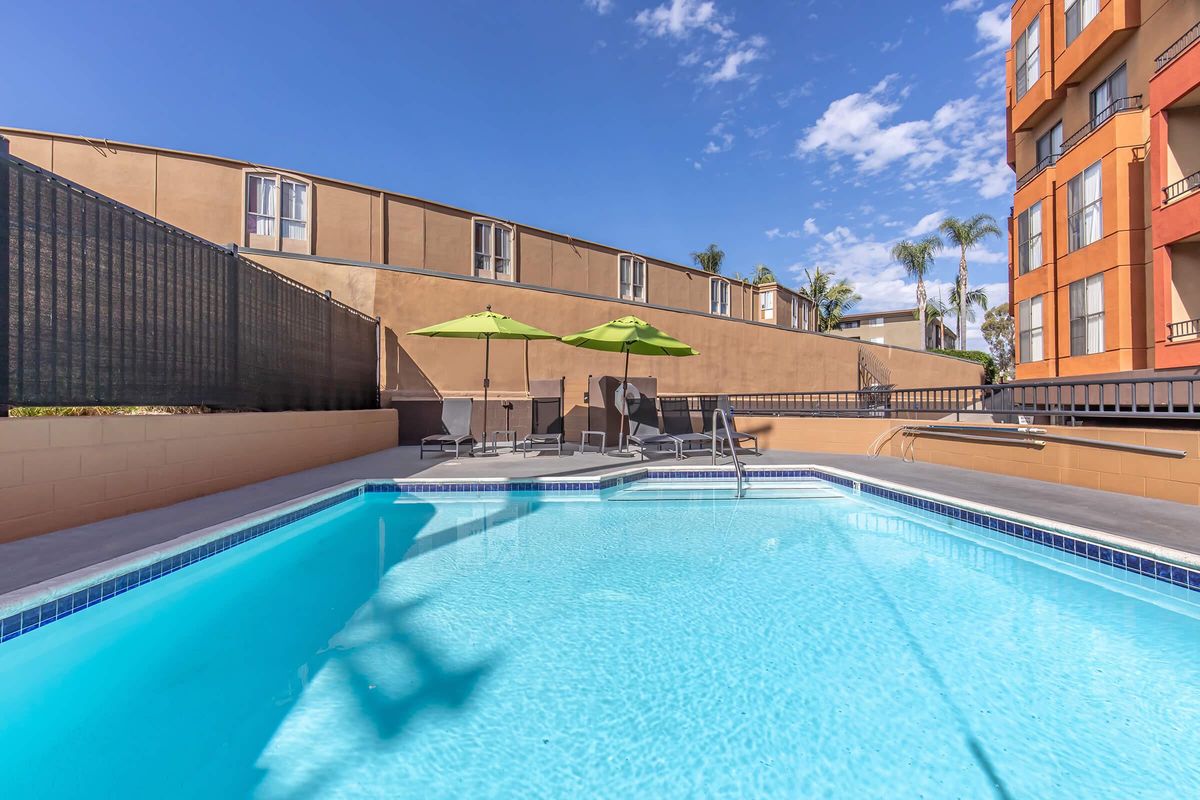 A clear blue swimming pool surrounded by a fence, with two green umbrellas and lounge chairs positioned on the pool deck. In the background, there are buildings and palm trees under a bright blue sky with a few clouds.