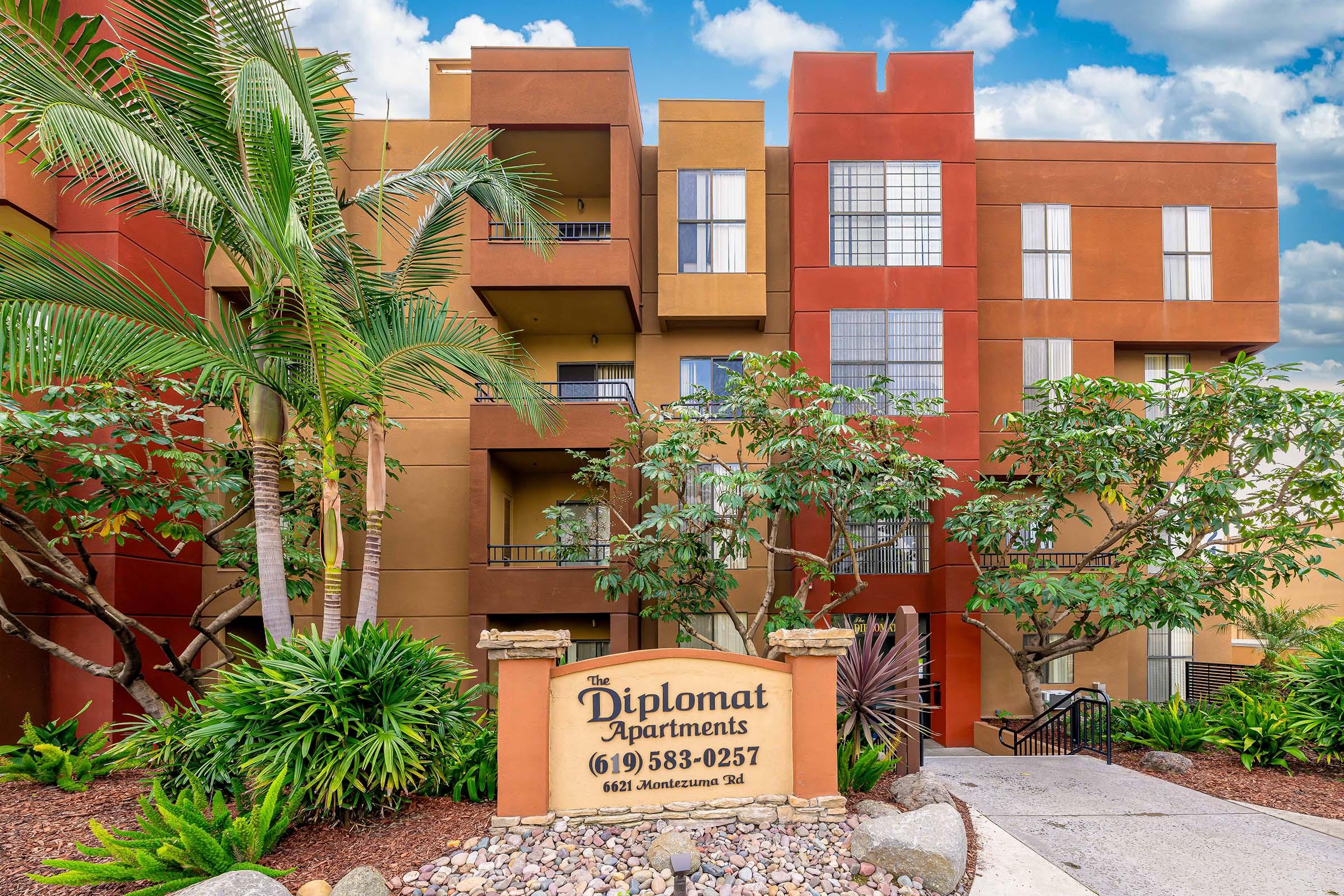 A colorful apartment building with the sign "The Diplomat Apartments" prominently displayed. The architecture features a mix of red and brown hues, surrounded by lush greenery and palm trees. The scene is set under a partly cloudy sky, showcasing a vibrant and inviting residential environment.
