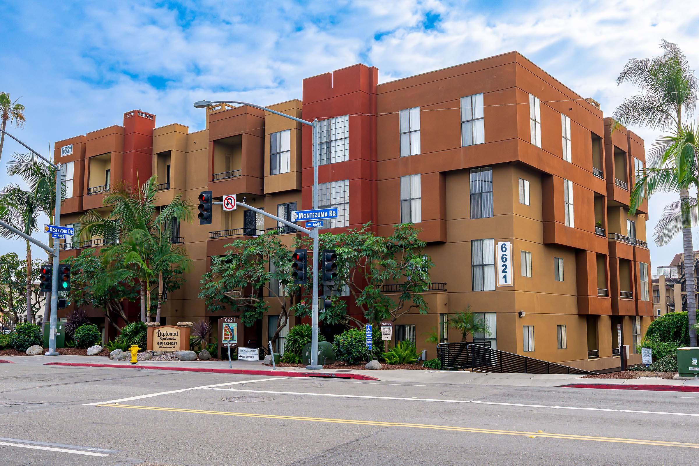 A modern multi-story apartment building in warm colors, located at the intersection of two streets. The building features large windows, palm trees, and signage indicating the address as 6621. Traffic lights and street signs are visible, suggesting a busy urban environment.