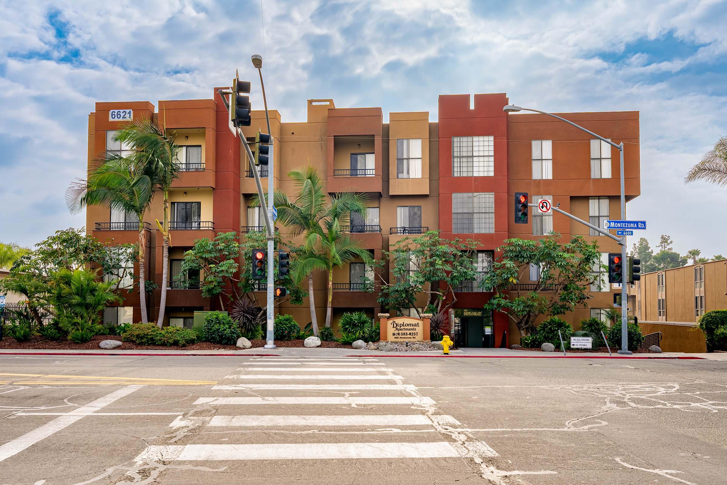 A modern multi-story building with a combination of red and orange exterior walls. It features balconies and large windows, surrounded by palm trees and landscaping. A street sign is visible nearby, along with traffic lights and a crosswalk in the foreground, indicating an urban area.