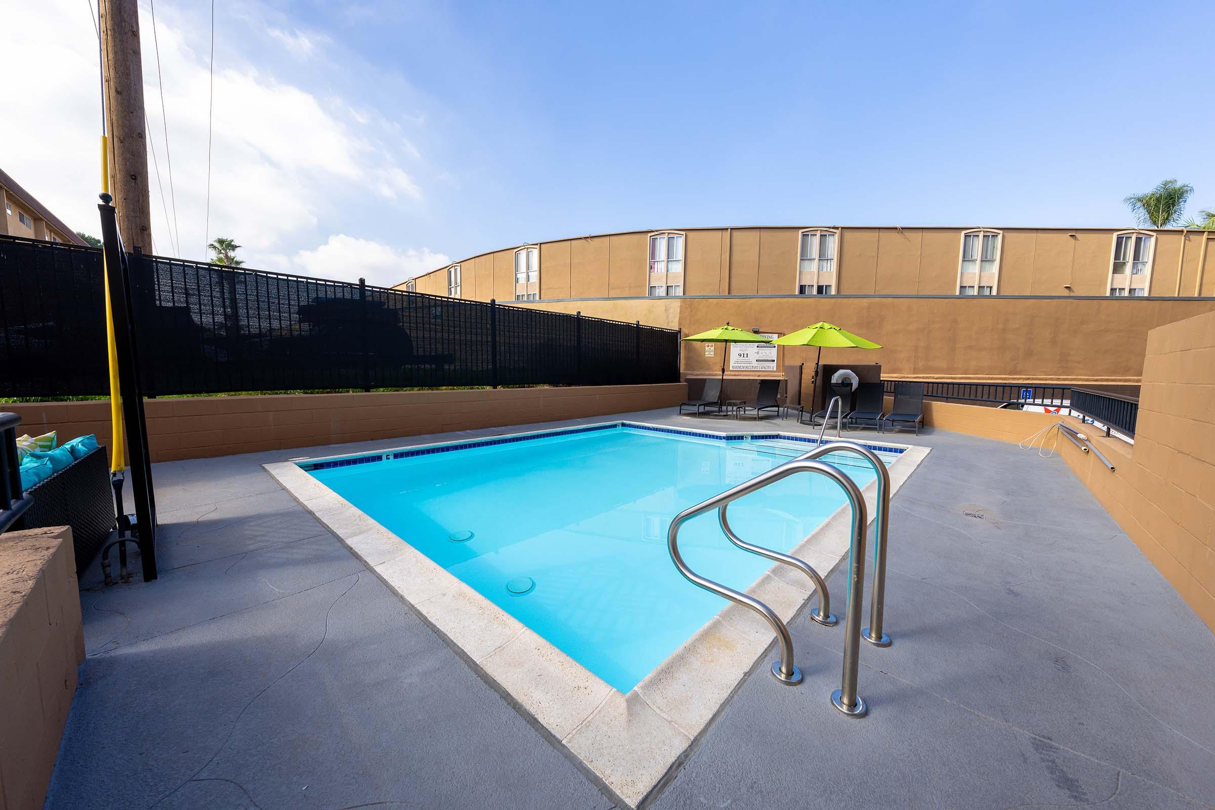 A clear swimming pool surrounded by a concrete deck, featuring a handrail for easy access. Sun loungers are positioned nearby under bright green umbrellas, with a backdrop of a beige building and a clear blue sky. The area is enclosed by a black fence, creating a private atmosphere.