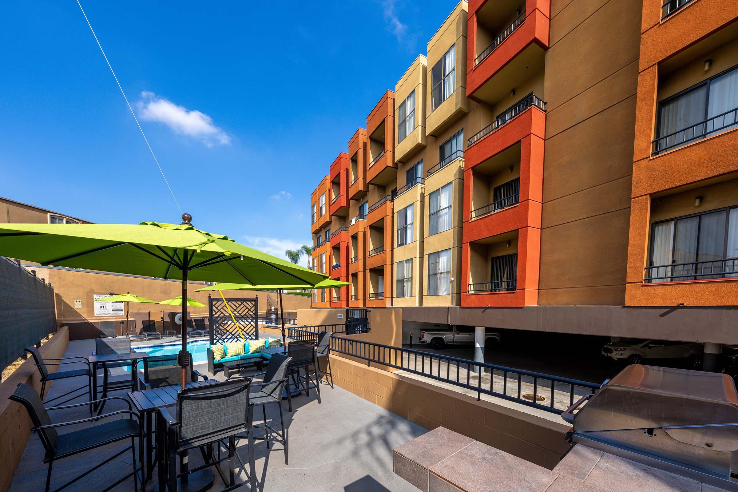 View of a hotel courtyard featuring a swimming pool surrounded by seating areas. Colorful umbrellas provide shade, and a multi-storied building with balconies stands in the background against a clear blue sky.