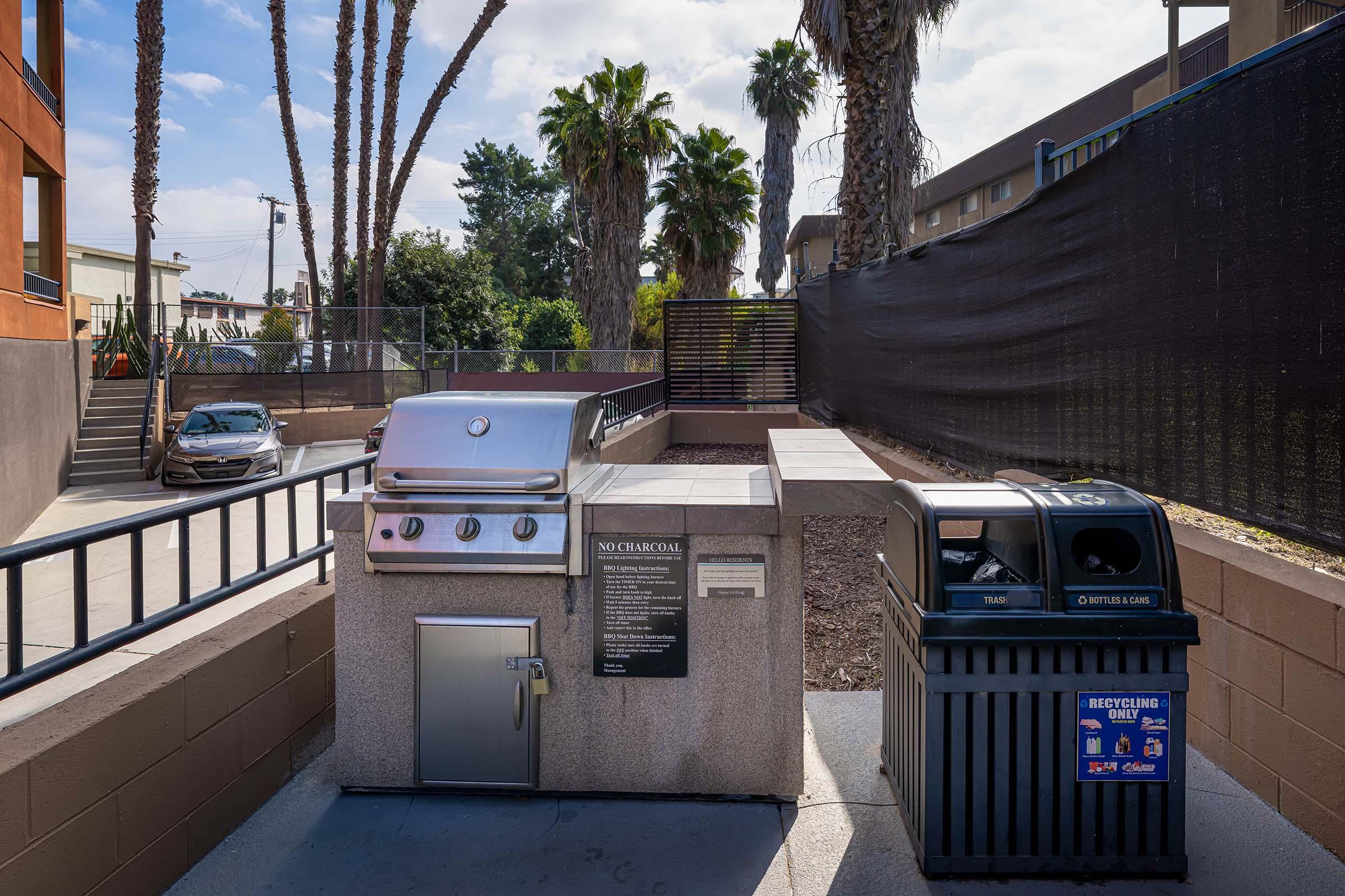 An outdoor grilling area featuring a modern stainless steel barbecue grill and a trash/recycling bin, surrounded by palm trees and a landscaped space in an urban setting. A car is parked nearby, and a privacy screen is visible in the background.