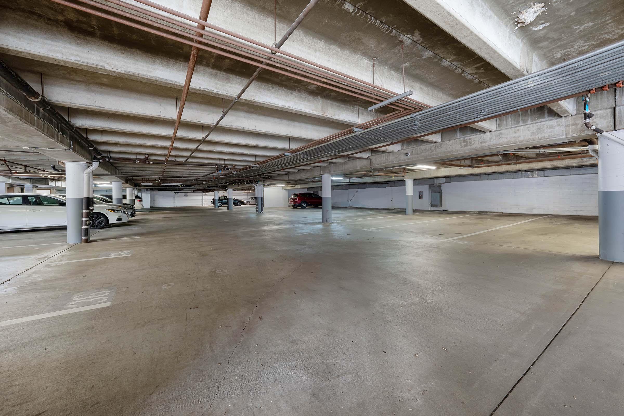 A spacious underground parking garage with concrete floors and exposed beams. Several parked cars are visible, including a white car on the left and a red car further back. The area is well-lit with columns supporting the ceiling and no other visible occupants.