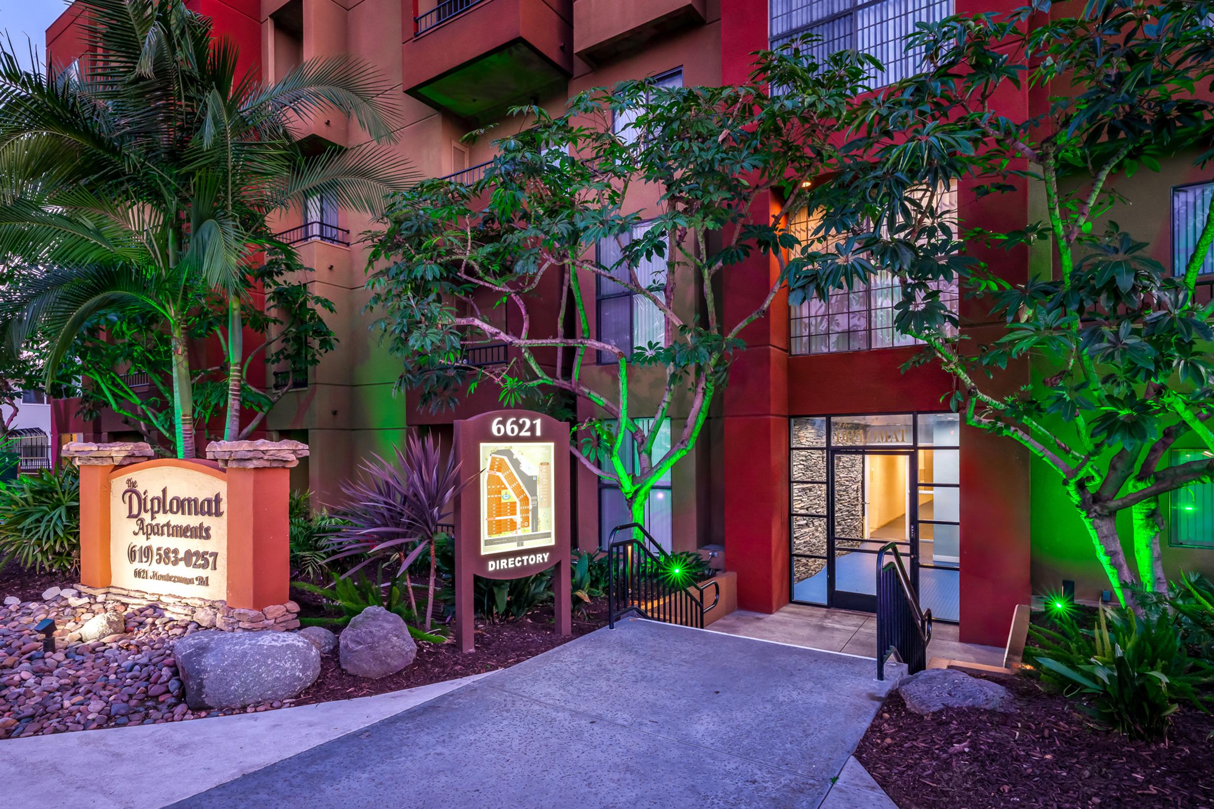 Entrance to the Diplomat Apartments, featuring a stone sign with the address "6621" and a directory. Lush greenery and palm trees surround the pathway, with the building's exterior illuminated by colorful lights, creating an inviting atmosphere.