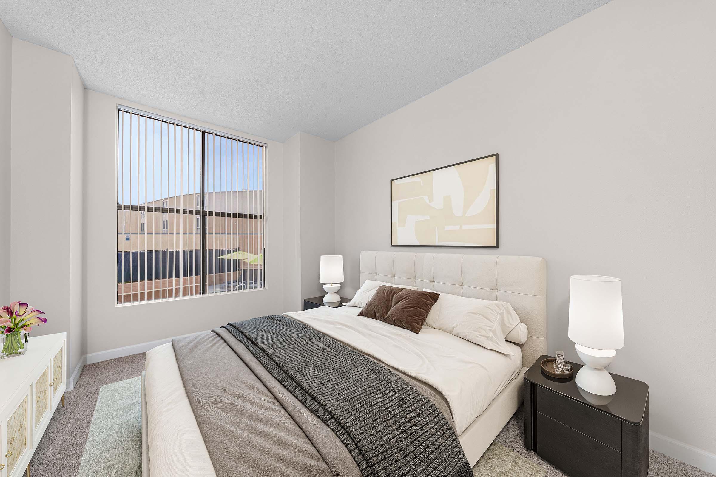 A cozy bedroom featuring a large bed with white linens and decorative pillows, a nightstand with a lamp, and a window with vertical blinds allowing natural light. There is a piece of abstract wall art above the bed and a small bouquet of flowers on the nightstand, adding a welcoming touch to the space.