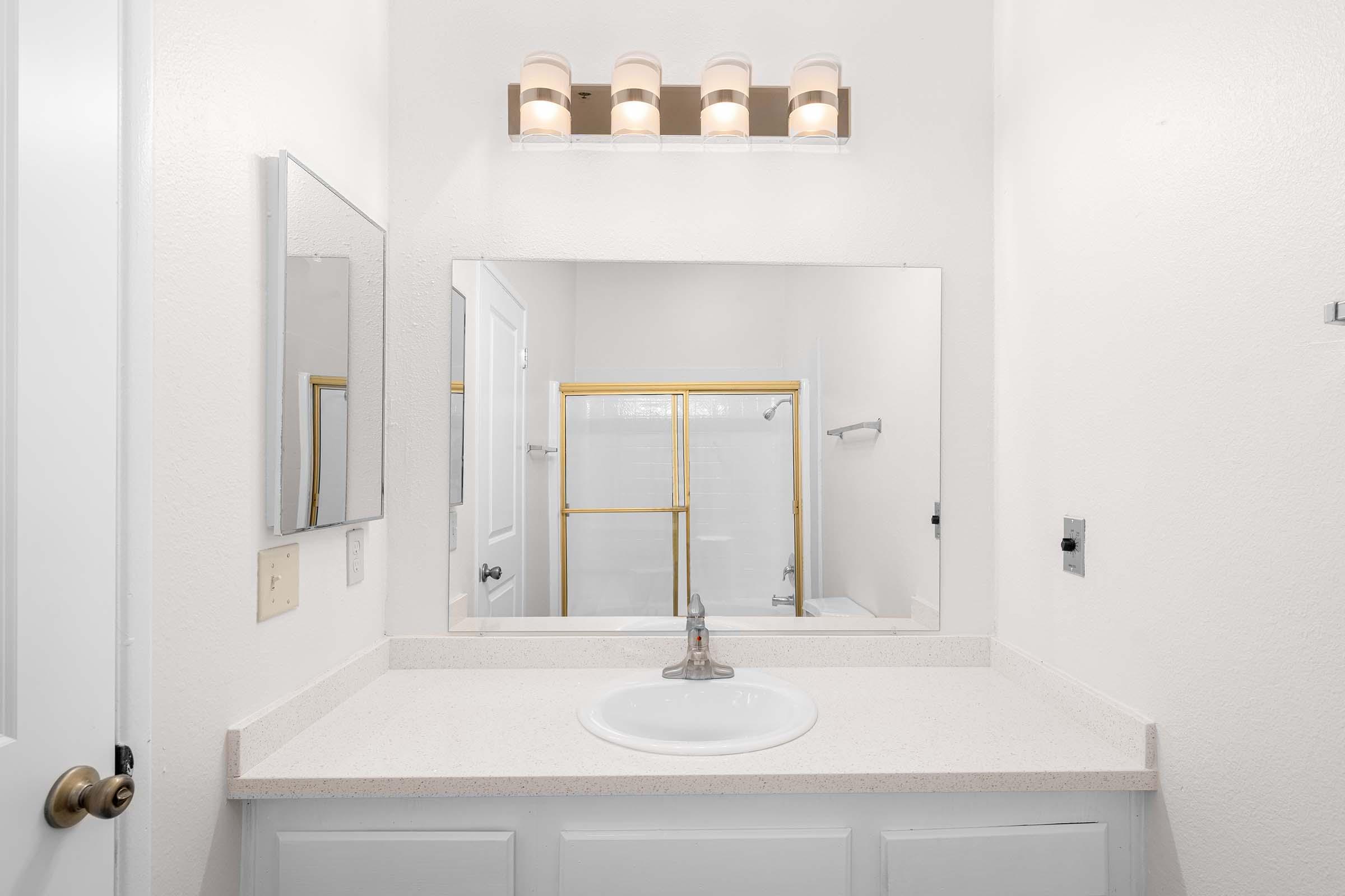 A clean bathroom with a white countertop, a sink, and a rectangular mirror above it. A light fixture with three bulbs is mounted above the mirror. The bathroom has a neutral color scheme, with white walls and a glimpse of a shower area with golden accents visible in the background.