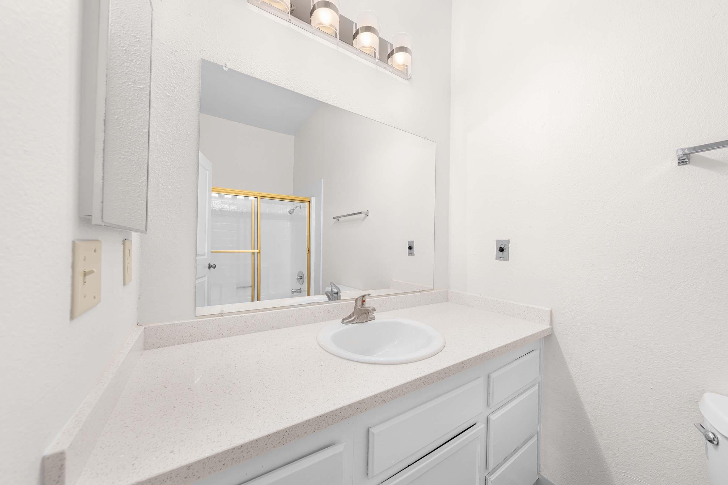 A clean, bright bathroom featuring a white countertop with a sink and faucet, a large mirror above, and minimalistic white cabinetry. The walls are painted in a light shade, and there’s a gold-framed shower enclosure visible in the background. Soft lighting fixtures are mounted above the mirror.