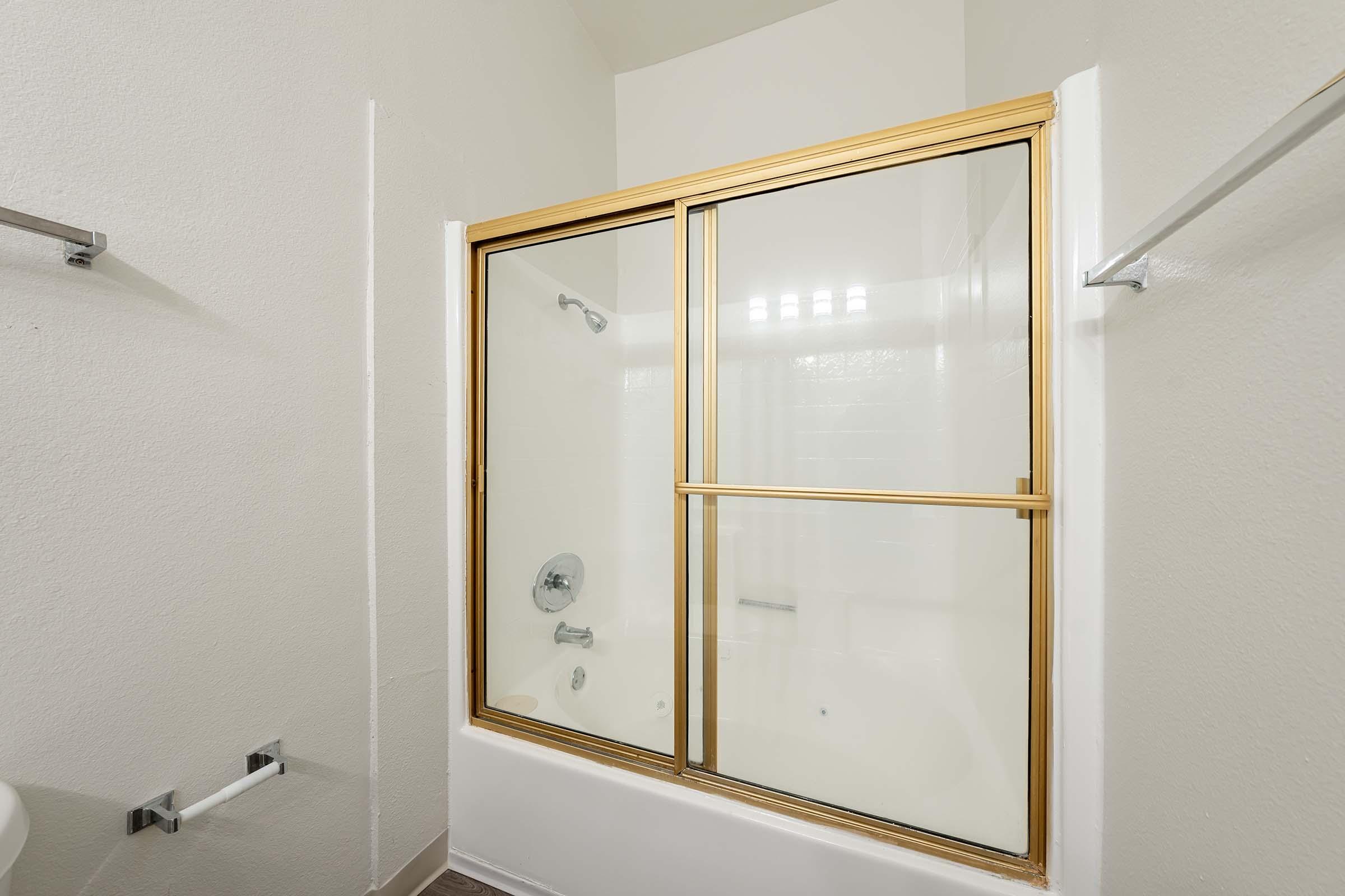 A clean and modern bathroom featuring a glass shower enclosure with gold trim. The walls are painted white, and the flooring is a light-colored tile. A towel rack is mounted on the wall, and there is a bathtub visible on the left side. The overall design is minimalist and bright.