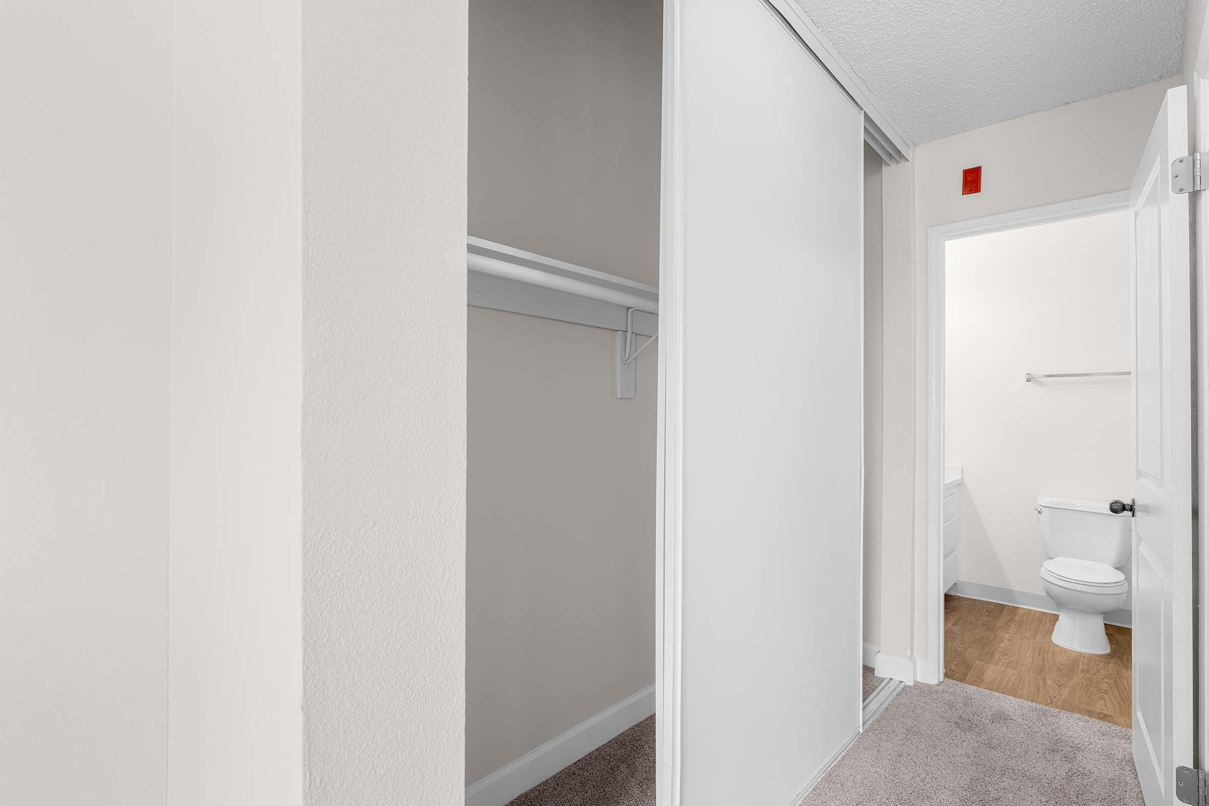 A clean, well-lit interior hallway featuring a sliding mirror door closet on the left with a shelf, and a bathroom doorway on the right. The bathroom is partially visible, with light-colored walls and a wooden floor. The space has a neutral color palette, giving it a modern and inviting feel.