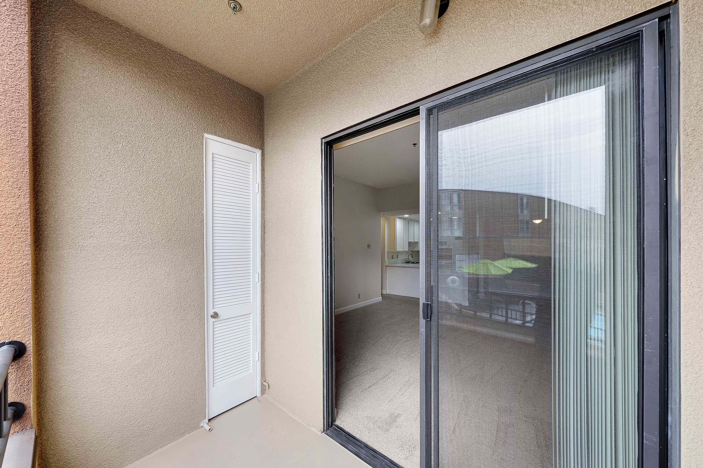 A view from a balcony showing a sliding glass door leading into an interior space with light-colored walls and a partially visible kitchen area. The floor is carpeted, and there is a door on the left side that leads to another room. Natural light is coming from the balcony.