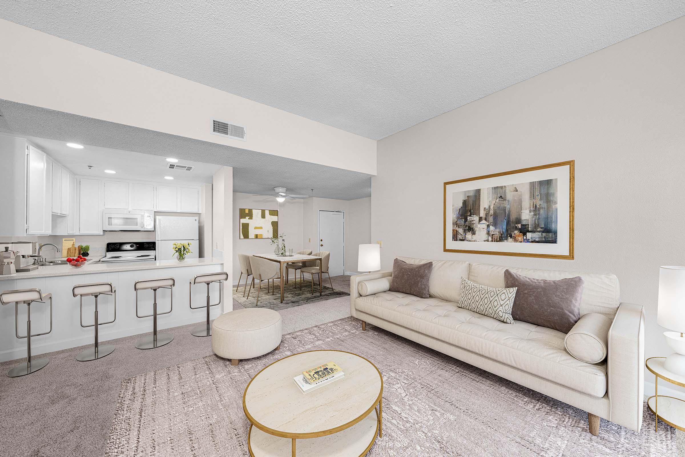 A modern living room featuring a light-colored sofa with plush cushions, a round coffee table, and a small ottoman. In the background, a dining area with a table and chairs is visible. The kitchen has white cabinets and appliances. Soft lighting and neutral tones create a cozy atmosphere.