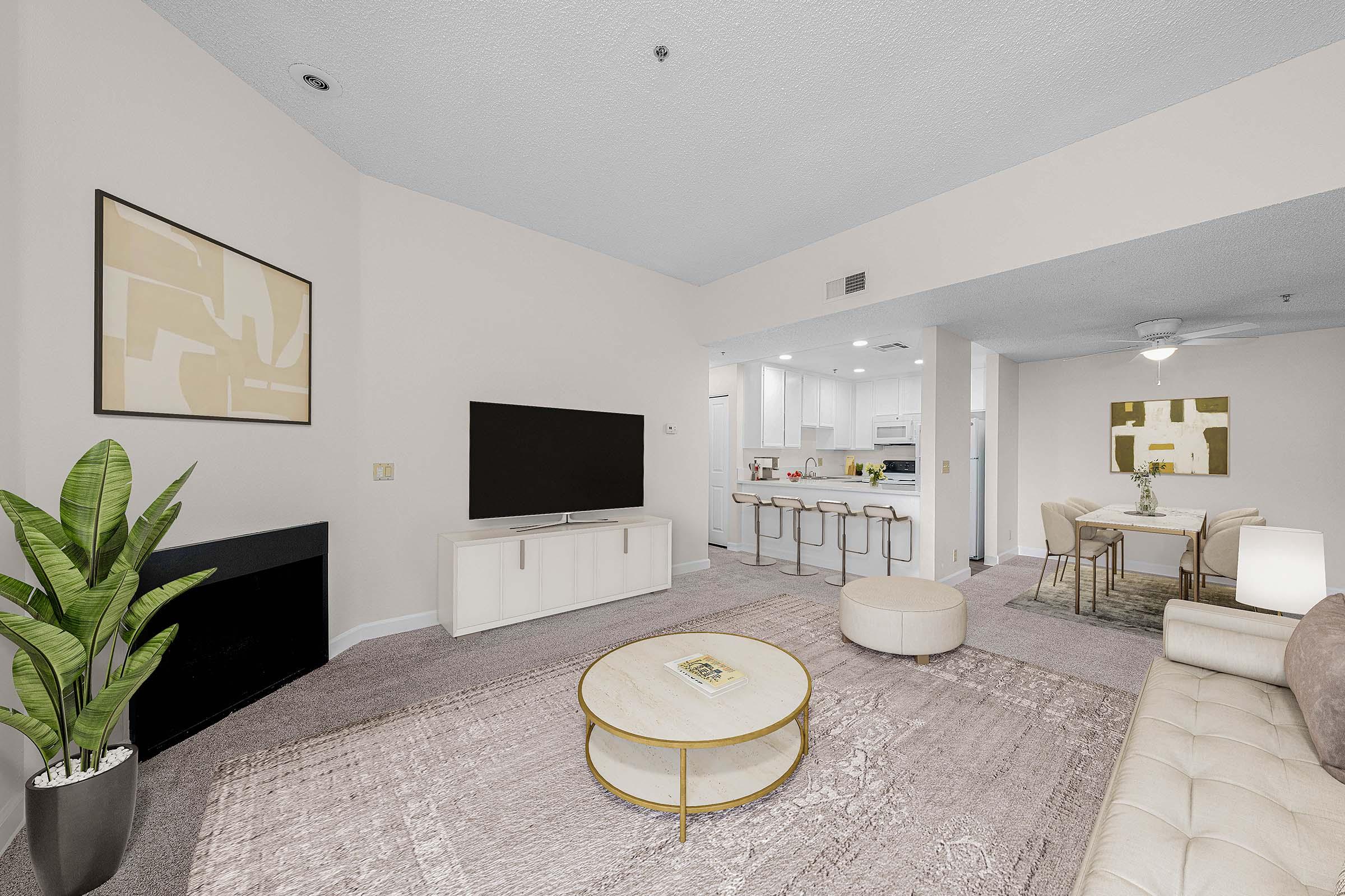 A modern living room featuring a beige sofa, circular coffee table, and a large flat-screen TV mounted on the wall. A potted plant adds greenery, while a kitchen area is visible in the background. The space has light-colored walls and a soft rug, creating a cozy and inviting atmosphere.
