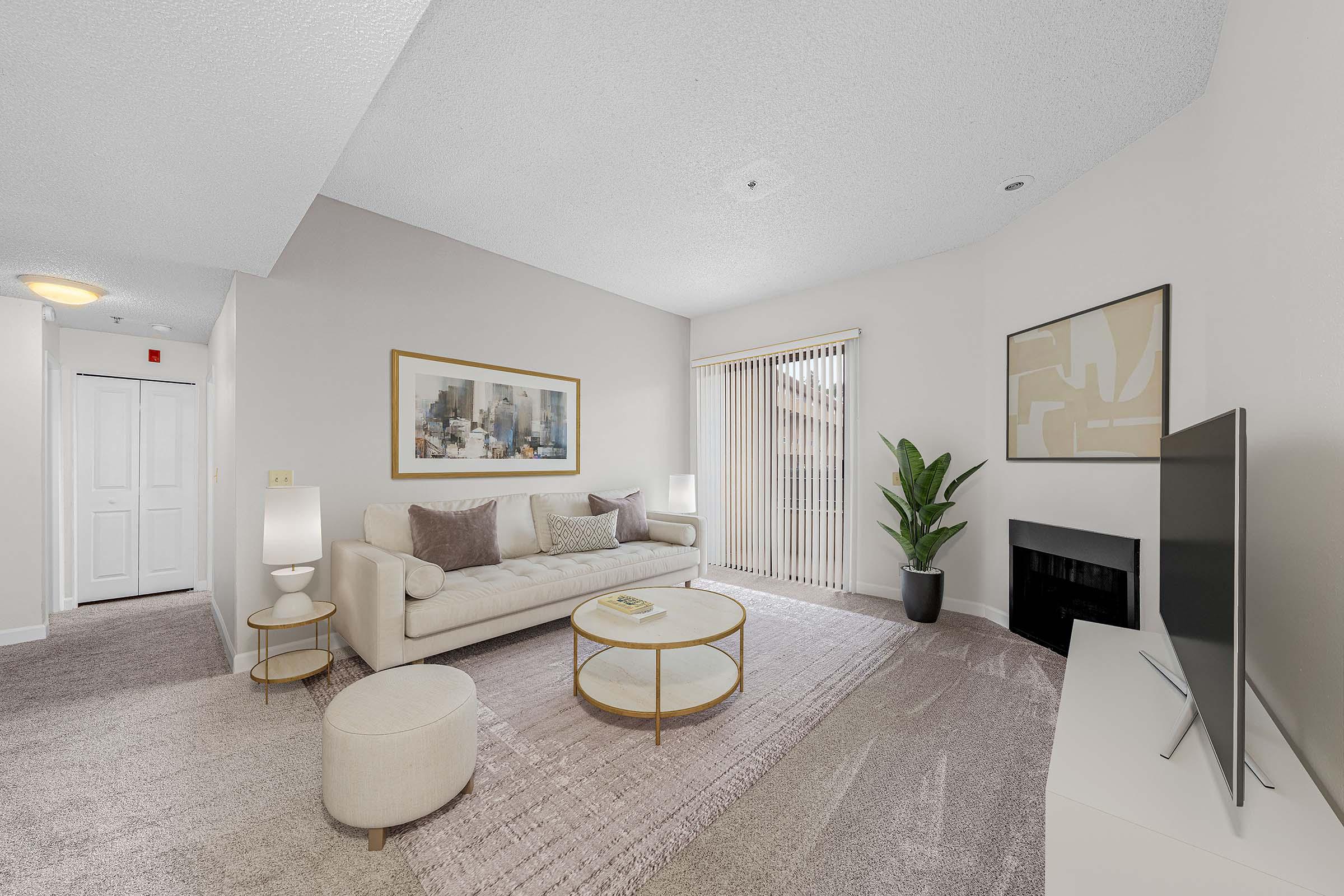 A modern living room featuring a light-colored sofa with decorative pillows, a round coffee table, and a flat-screen TV on a white cabinet. There is a potted plant in the corner, a rug on the floor, and large windows with vertical blinds allowing natural light. Wall art adds a touch of elegance, and the space has a cozy atmosphere.