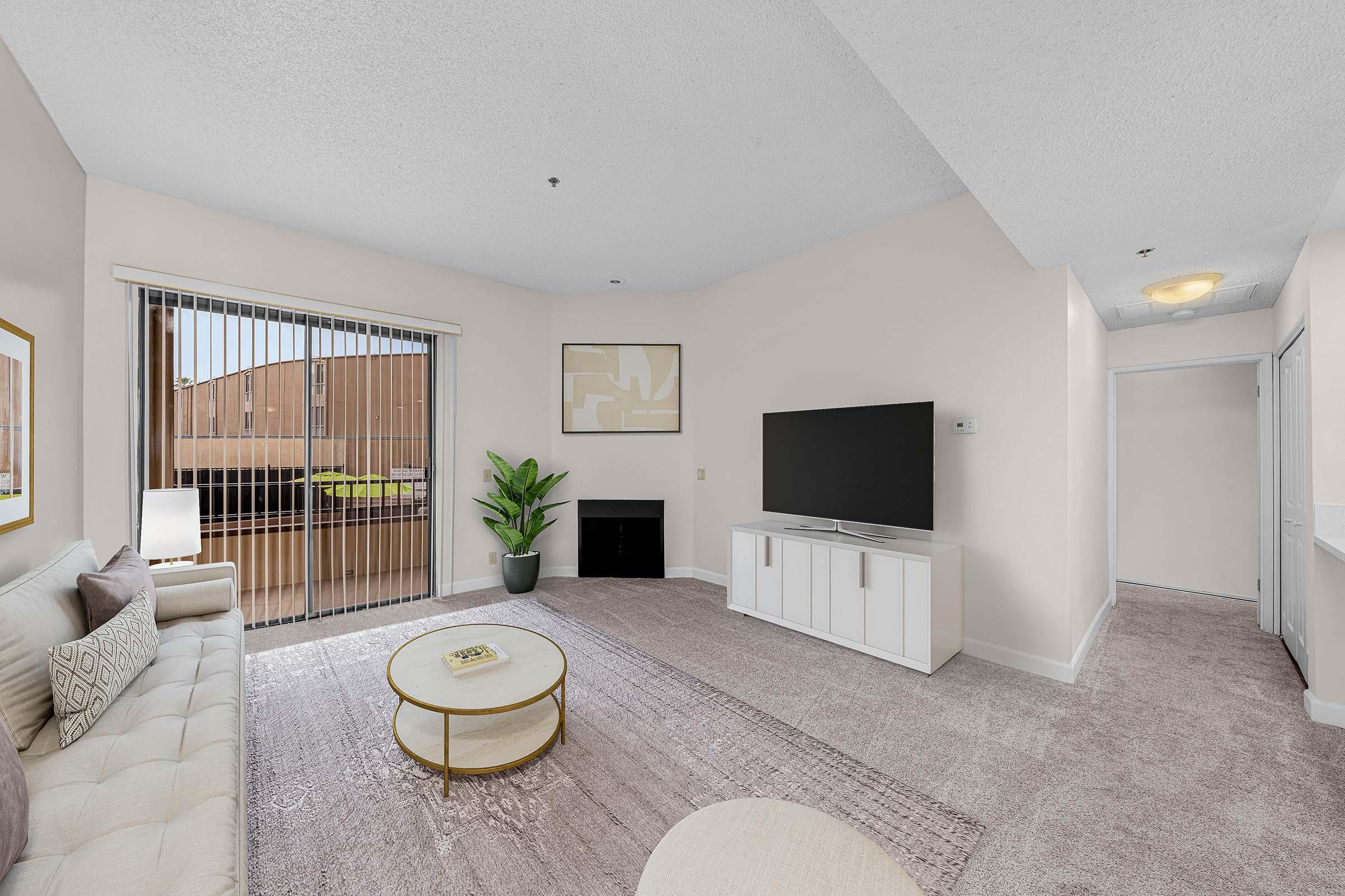 A modern living room featuring a light-colored couch and a round coffee table on a soft rug. A large flat-screen TV is mounted on a white stand. Large sliding glass doors provide natural light and a view of an outdoor space. Walls are painted in neutral tones, and a potted plant adds a touch of greenery.