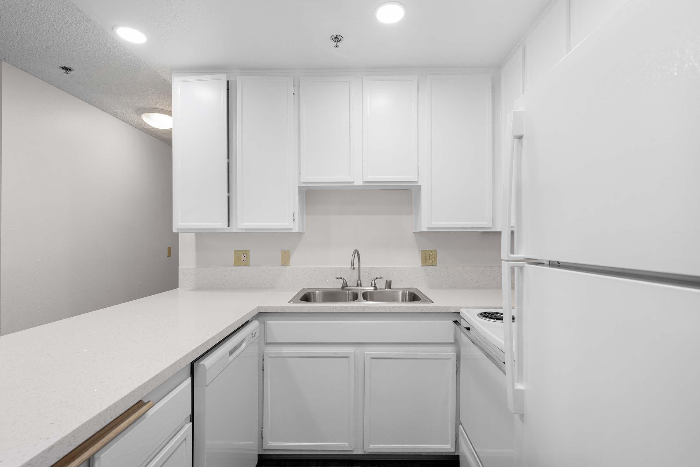 A modern kitchen featuring white cabinetry, a double sink, and a countertop with a smooth finish. There’s a white refrigerator and an electric stovetop, with a bright and clean aesthetic enhanced by natural light. The walls are neutral-colored, creating an open and welcoming space.