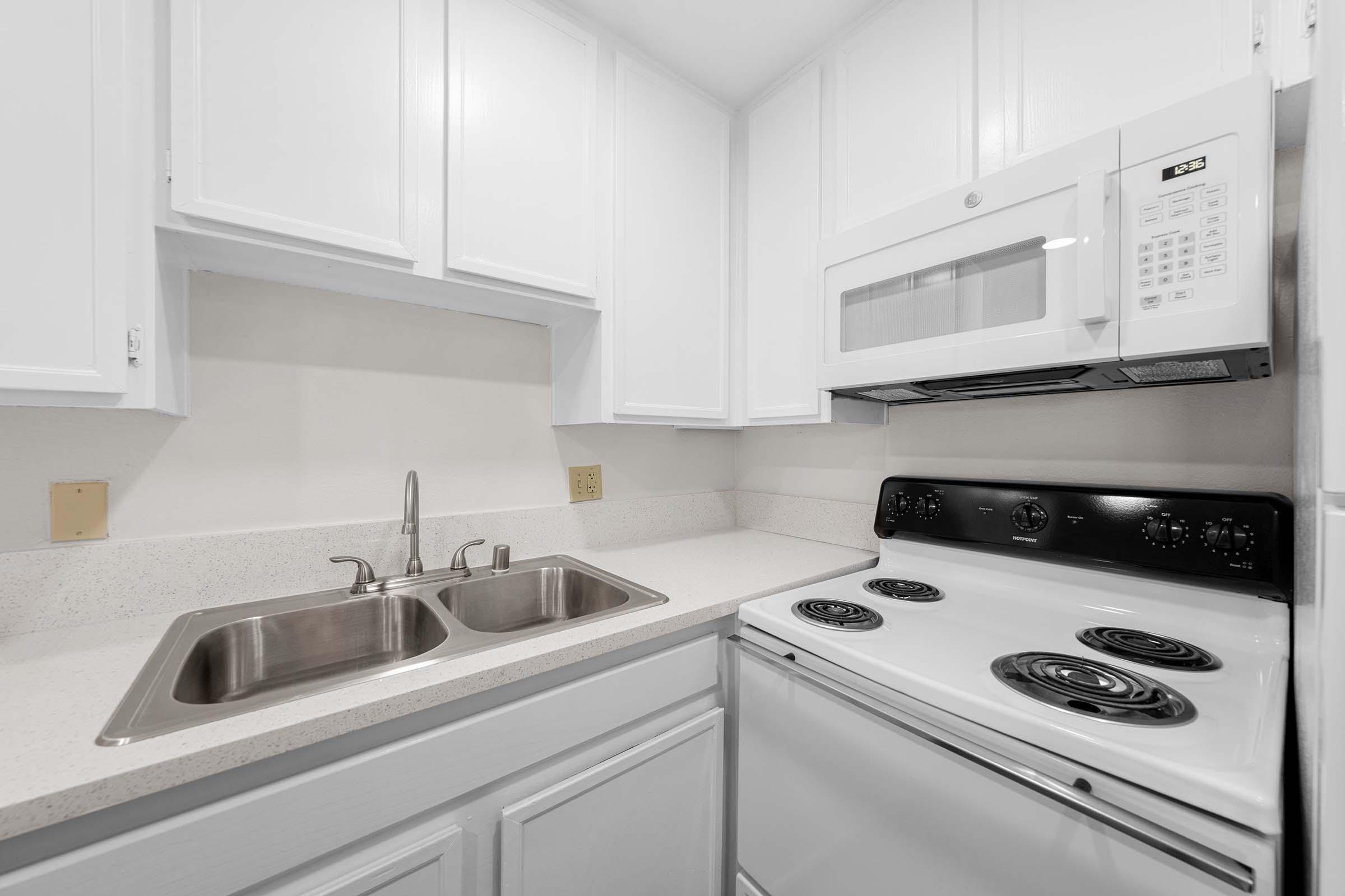 A modern kitchen featuring white cabinetry, a double sink, a microwave mounted above the stove, and a white stove with four burners. The countertop is light-colored, and the overall design is clean and contemporary, suitable for a small cooking space.