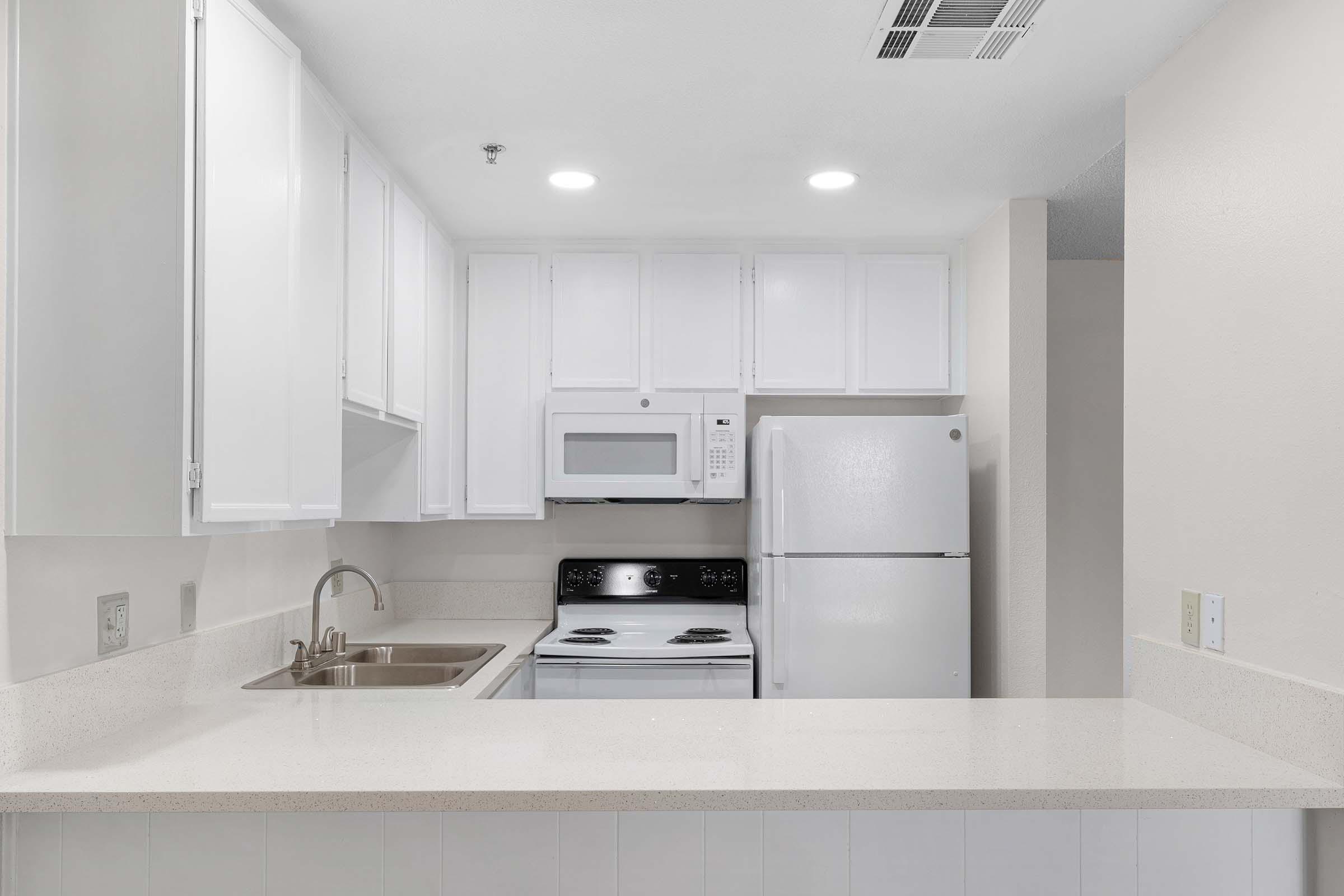 A modern kitchen featuring white cabinets, a stainless steel sink, a stove, a microwave, and a white refrigerator. The countertop is light-colored, and the space is well-lit with recessed ceiling lights, creating a bright and functional environment.