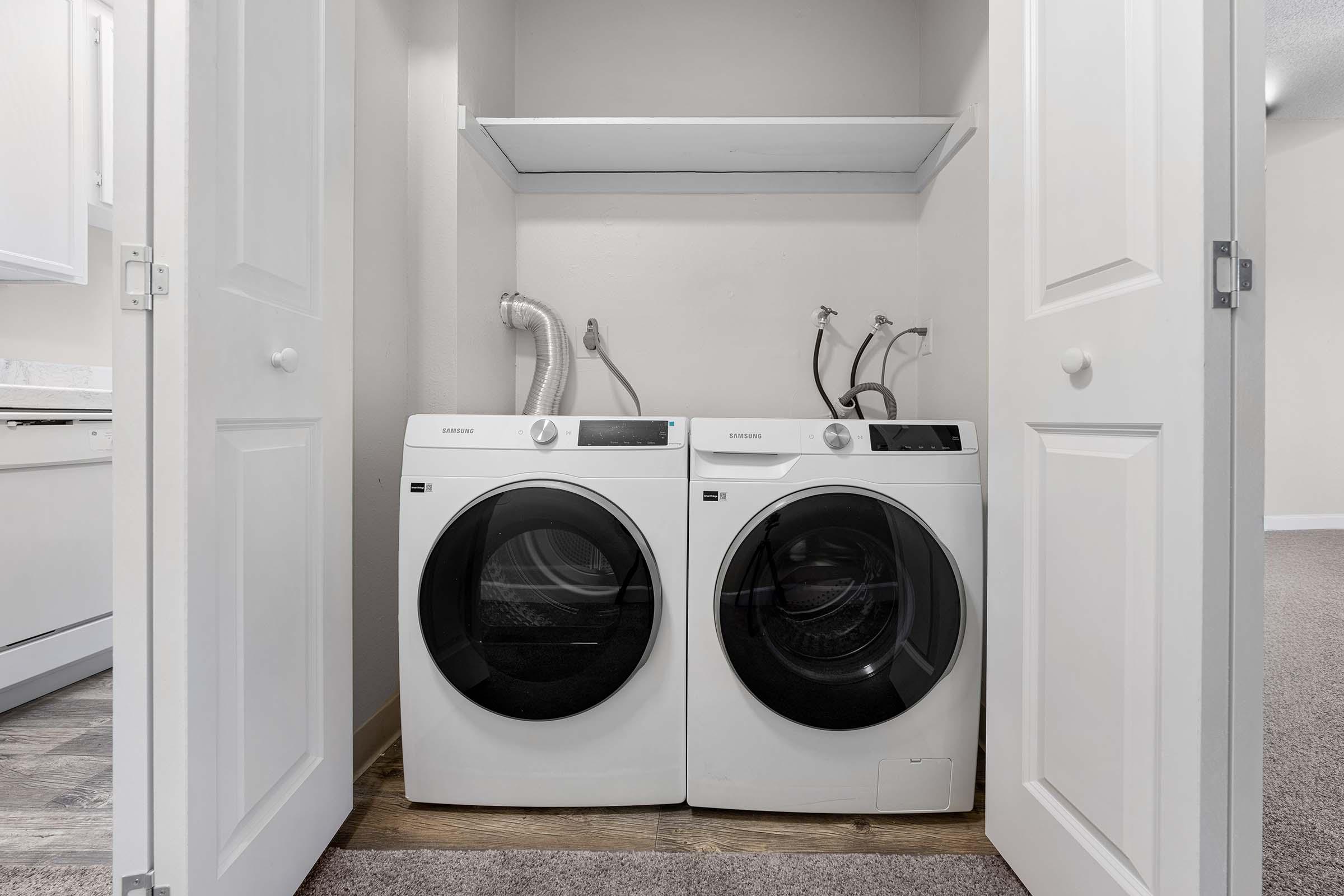 A compact laundry area featuring a white washer and dryer set side by side. The appliances are neatly stored in a closet space with double doors slightly open. There is a shelf above the machines and visible hoses connected to the back of the appliances. The floor has a carpeted finish.