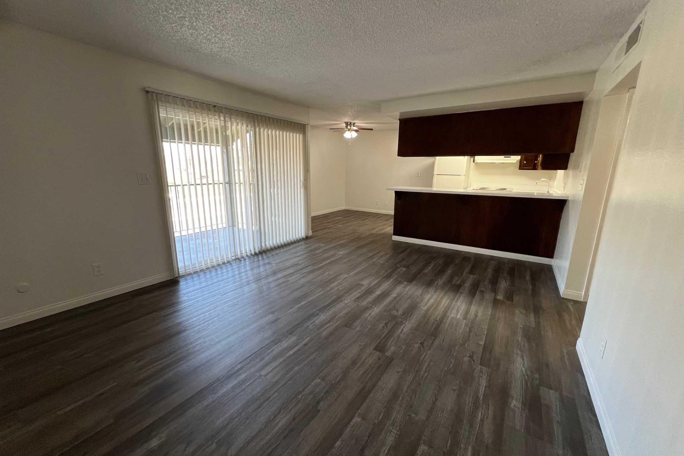 A spacious living area with wooden floors and white walls. There are sliding glass doors leading to a balcony, and a kitchen area with a bar counter visible in the background. A ceiling fan is installed in the room, providing a bright and open atmosphere.