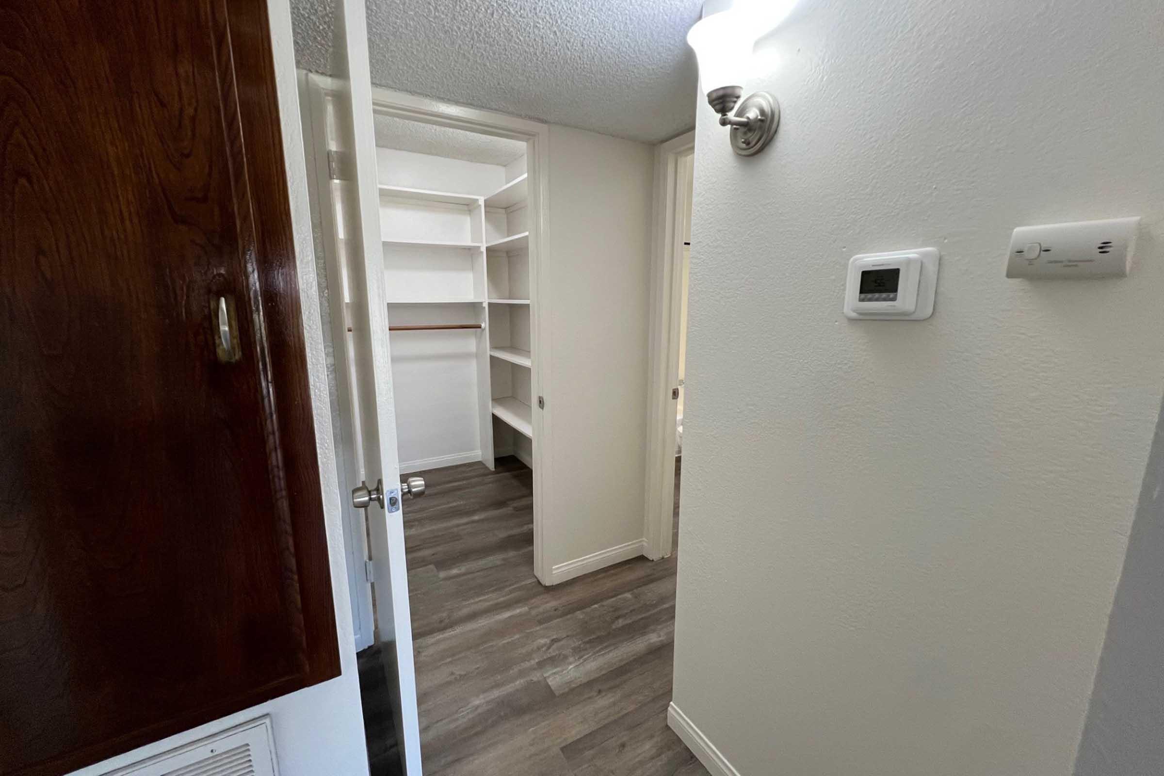 A narrow hallway leading to a walk-in closet with empty shelves on the left and a door on the right. The flooring is a modern wood-like design, and a wall-mounted light fixture is visible. A thermostat is also mounted on the wall near the door.