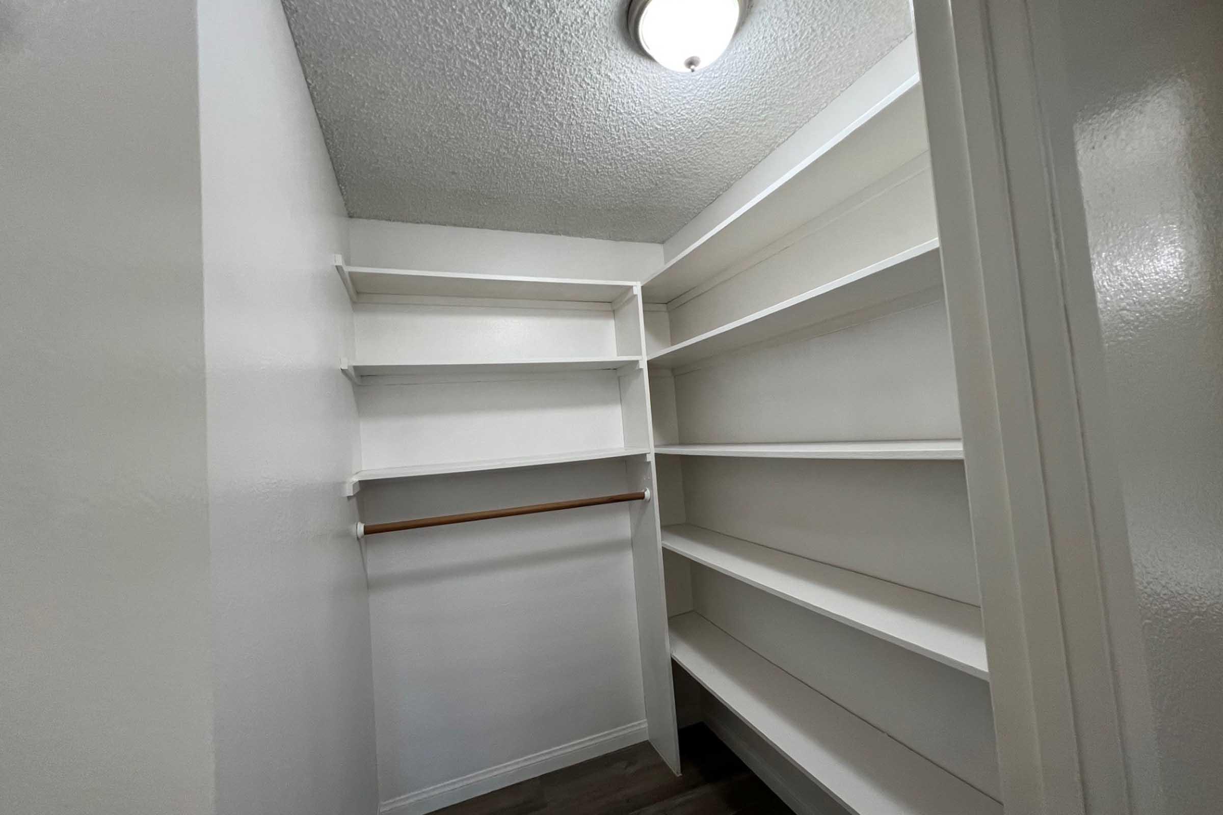 A well-lit, empty closet featuring white shelves and a single wooden rod for hanging clothes. The walls are painted white, and the ceiling has a simple light fixture. The floor is smooth, contributing to a clean and organized appearance.