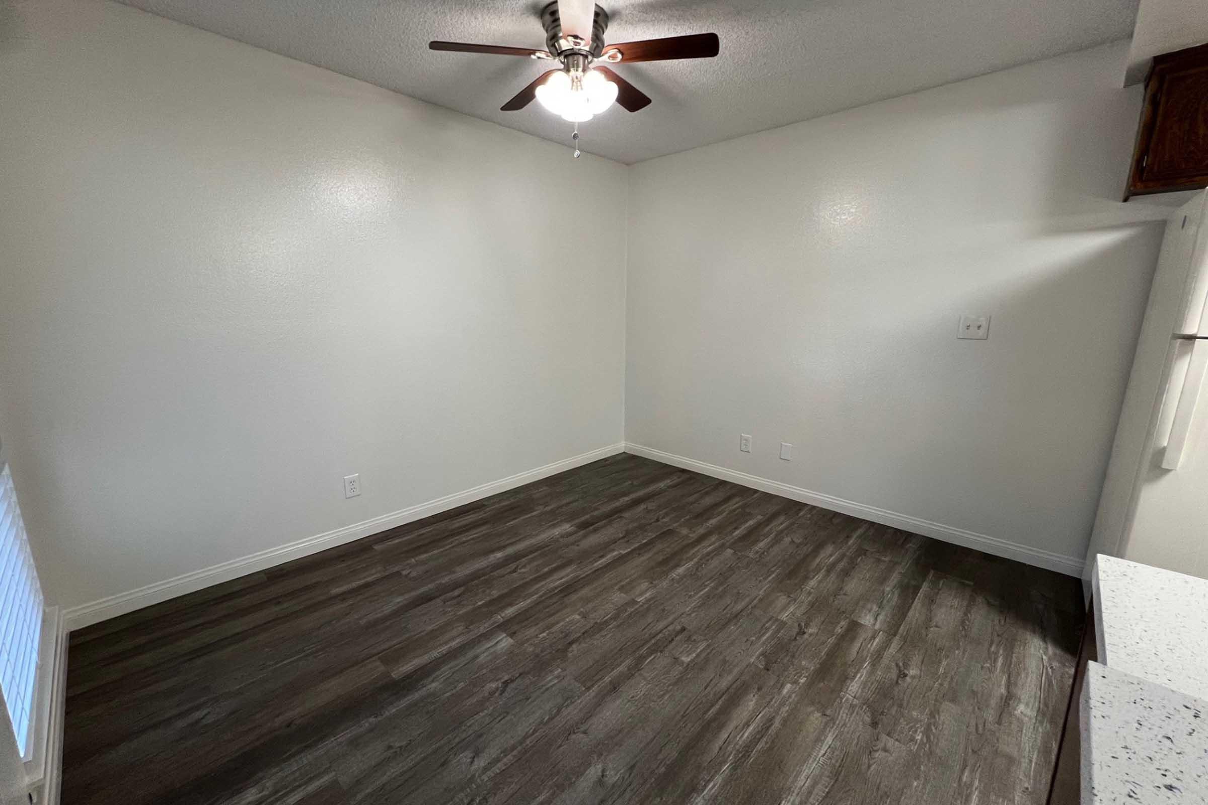 Empty room featuring light-colored walls, a ceiling fan with wooden blades, and a dark wood-like floor. There is a small section of cabinetry on the right side, and the room is well-lit. The space appears clean and ready for furnishings.
