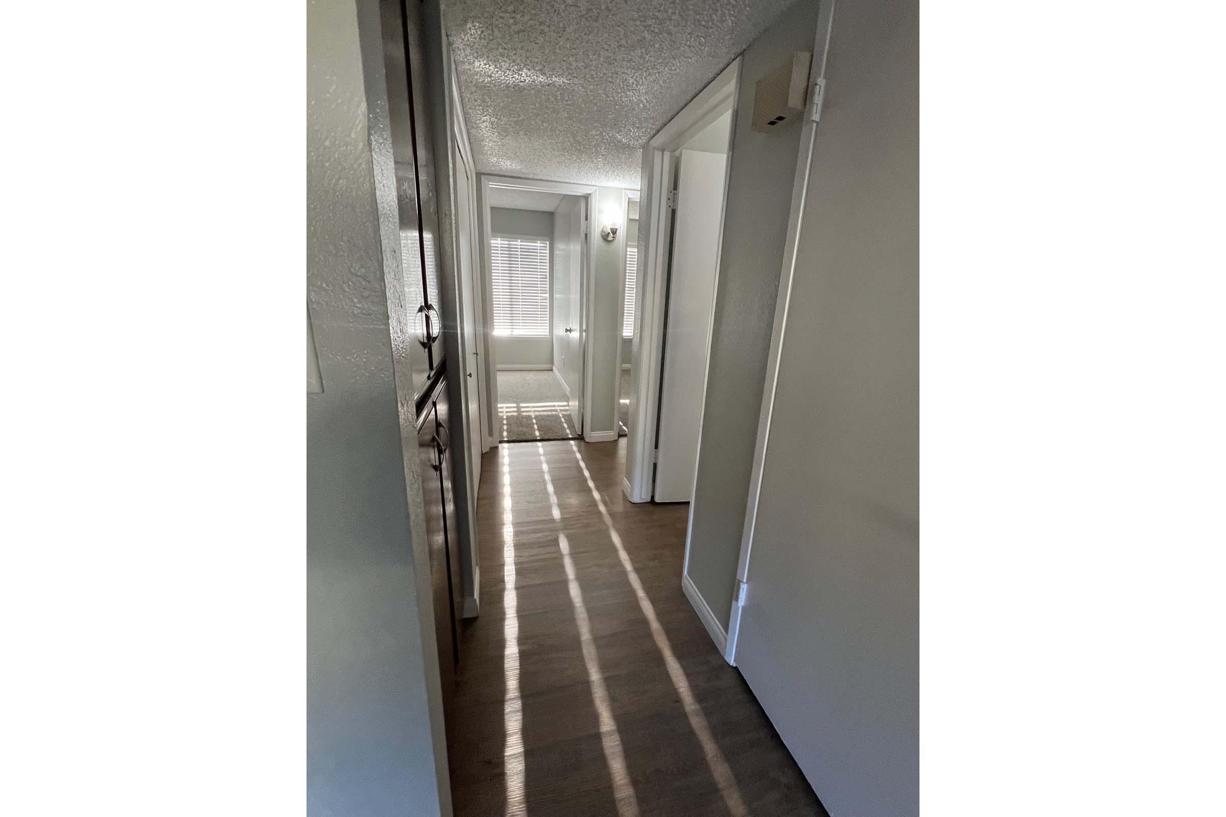 A well-lit hallway featuring light-colored walls and wooden flooring. The corridor extends toward a doorway at the end, with sunlight casting shadows on the floor. There are doors on either side, and the space has a clean and minimalist aesthetic.