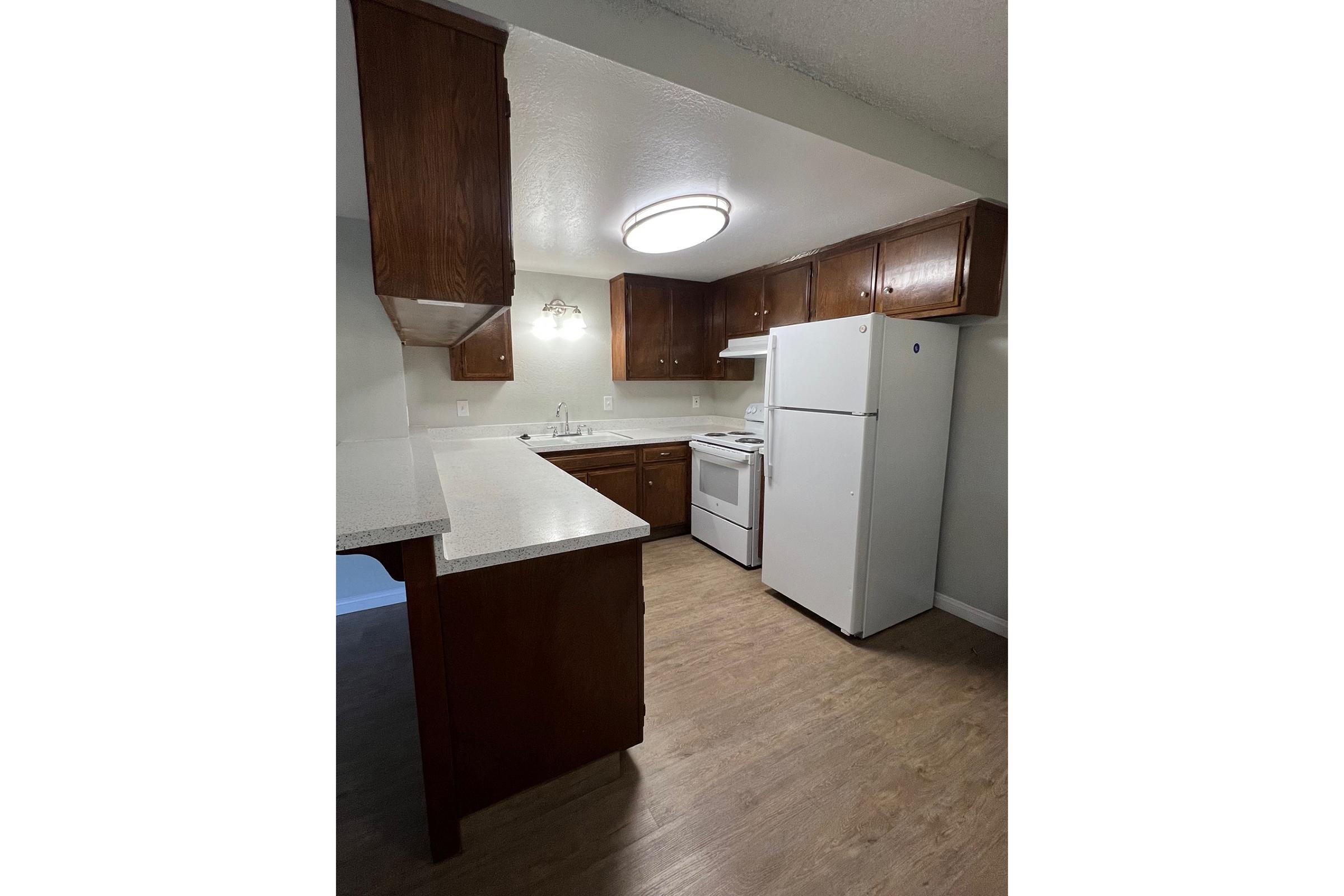 A view of a modern kitchen featuring brown wooden cabinets, a white refrigerator, and a white stove. The countertop is light-colored, and there is a round ceiling light providing illumination. The walls are painted a soft color, and the floor is a light wood laminate.
