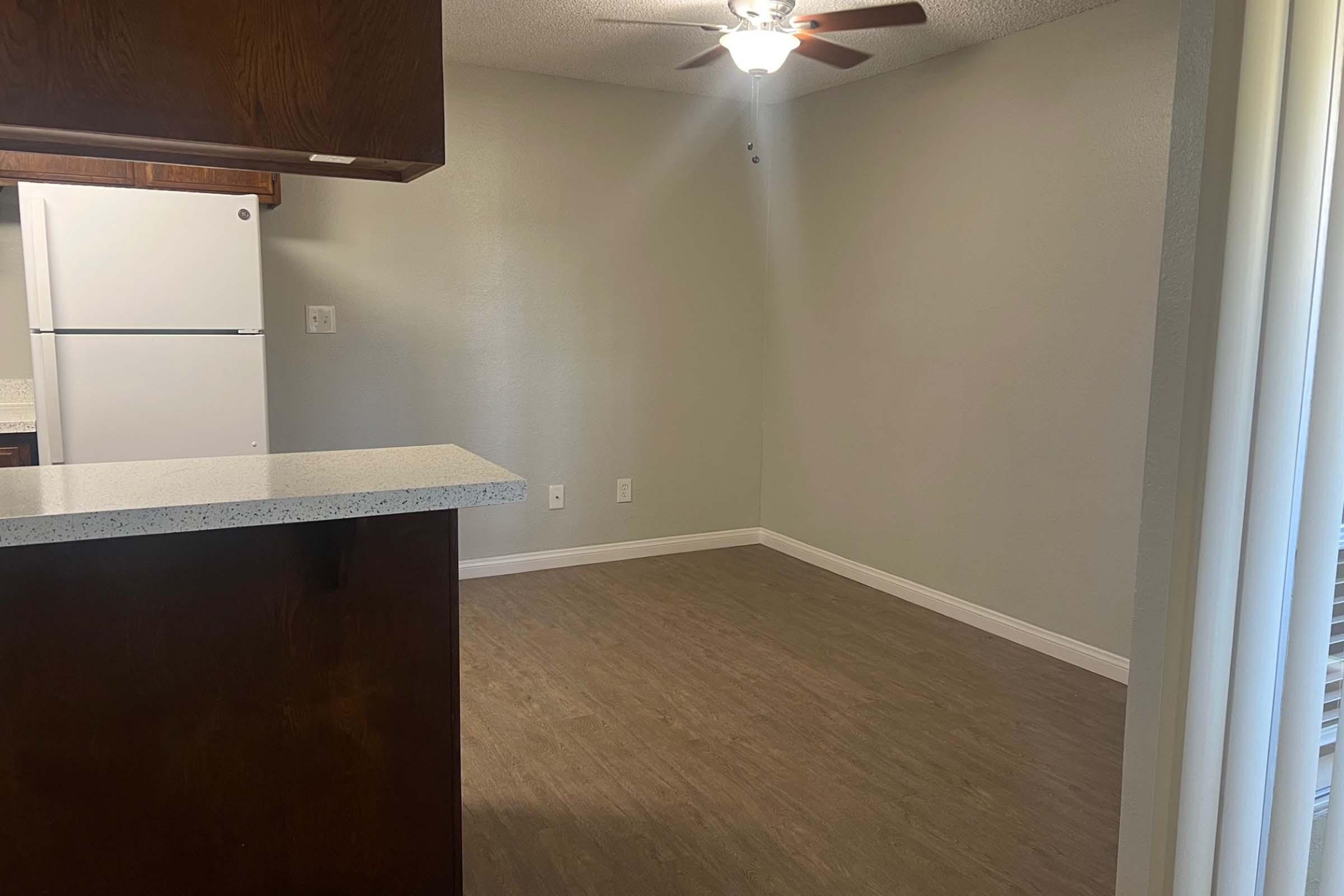 A spacious, empty room with light gray walls and a wooden floor. A ceiling fan hangs from the ceiling. On the left, a counter overlooks a small kitchen area with a white refrigerator. Natural light enters through a window, illuminating the space.