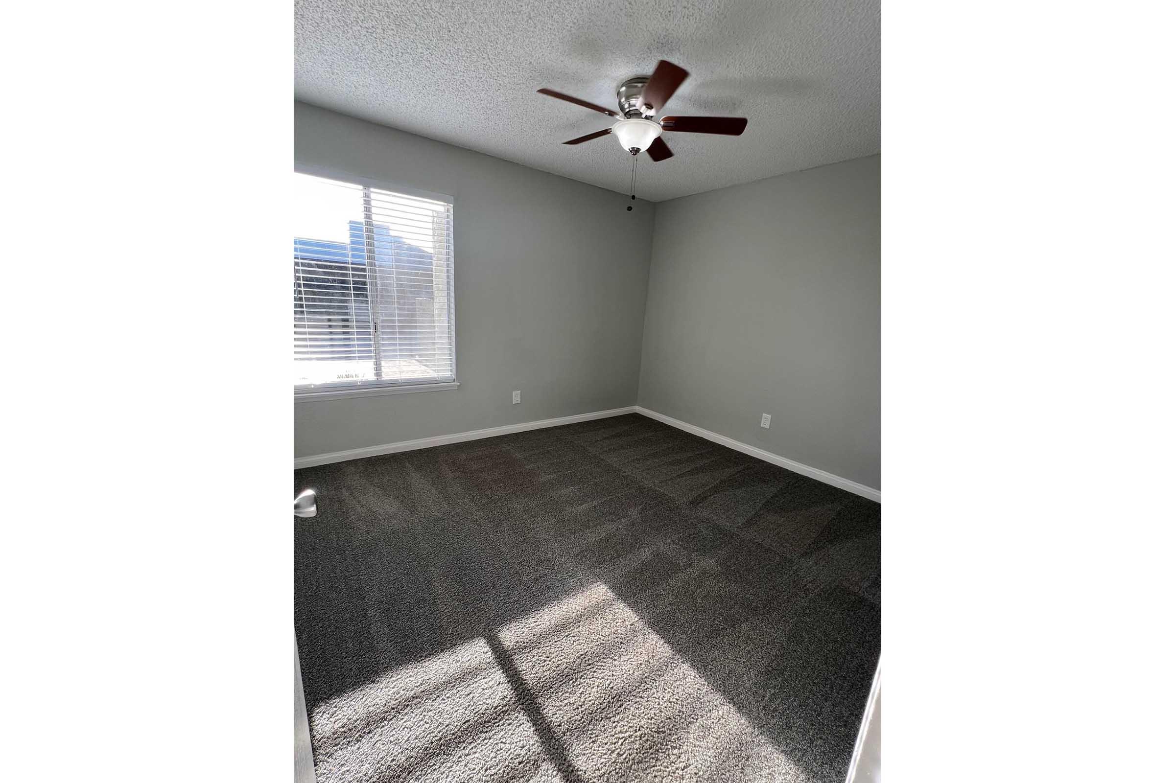 A well-lit, empty bedroom featuring light gray walls and a ceiling fan. The room has a window with blinds, allowing natural light to filter in, and is carpeted in a light gray shade. The interior appears clean and freshly painted, creating a blank canvas for potential furnishings and decor.