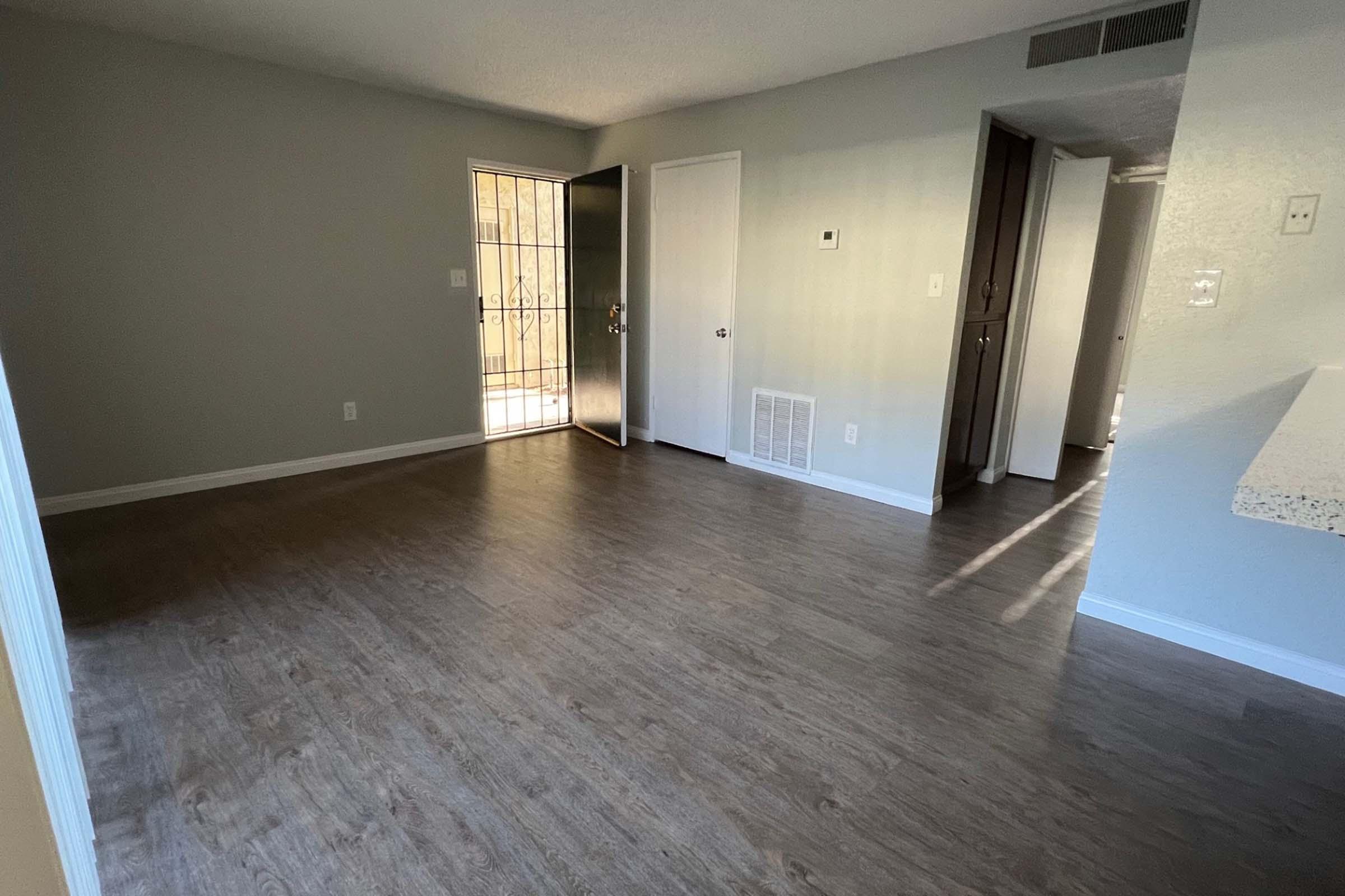 An empty room with light-colored walls and a wooden floor. There is a doorway leading to a balcony, a closed door on the right, and a wall with an air vent. The space is bright with natural light coming from the balcony door.