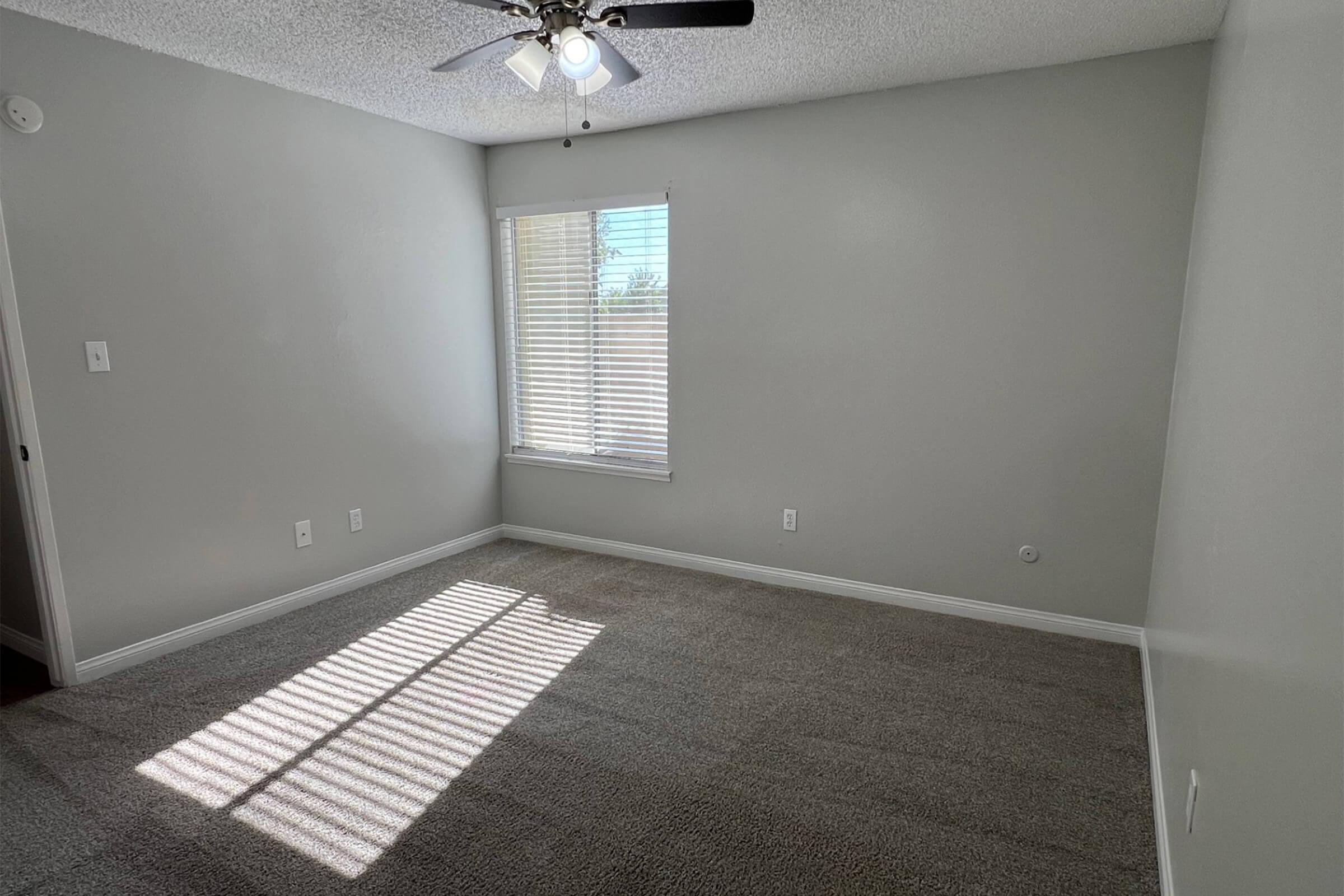 An empty room with beige carpet and light gray walls. A window with blinds allows natural light to enter, casting shadows on the floor. A ceiling fan is visible, and there are two wall switches on the left. The space appears clean and well-lit, suitable for various uses.