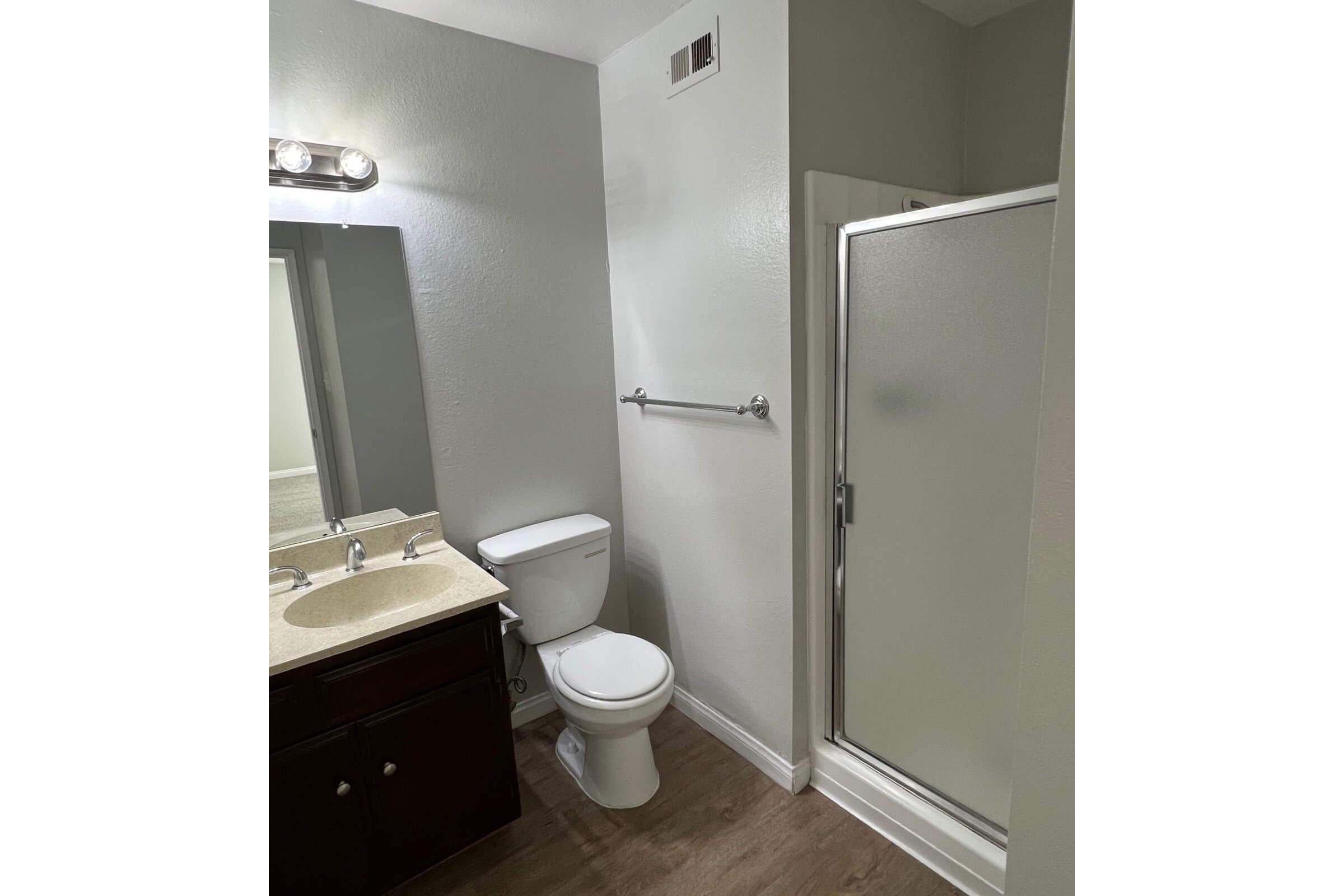 A modern bathroom featuring a white toilet, a glass shower enclosure, a double sink vanity with a light-colored countertop, and a large mirror. The walls are painted gray, and there is a towel bar near the shower. The flooring is a wood-like laminate, creating a clean and contemporary look.