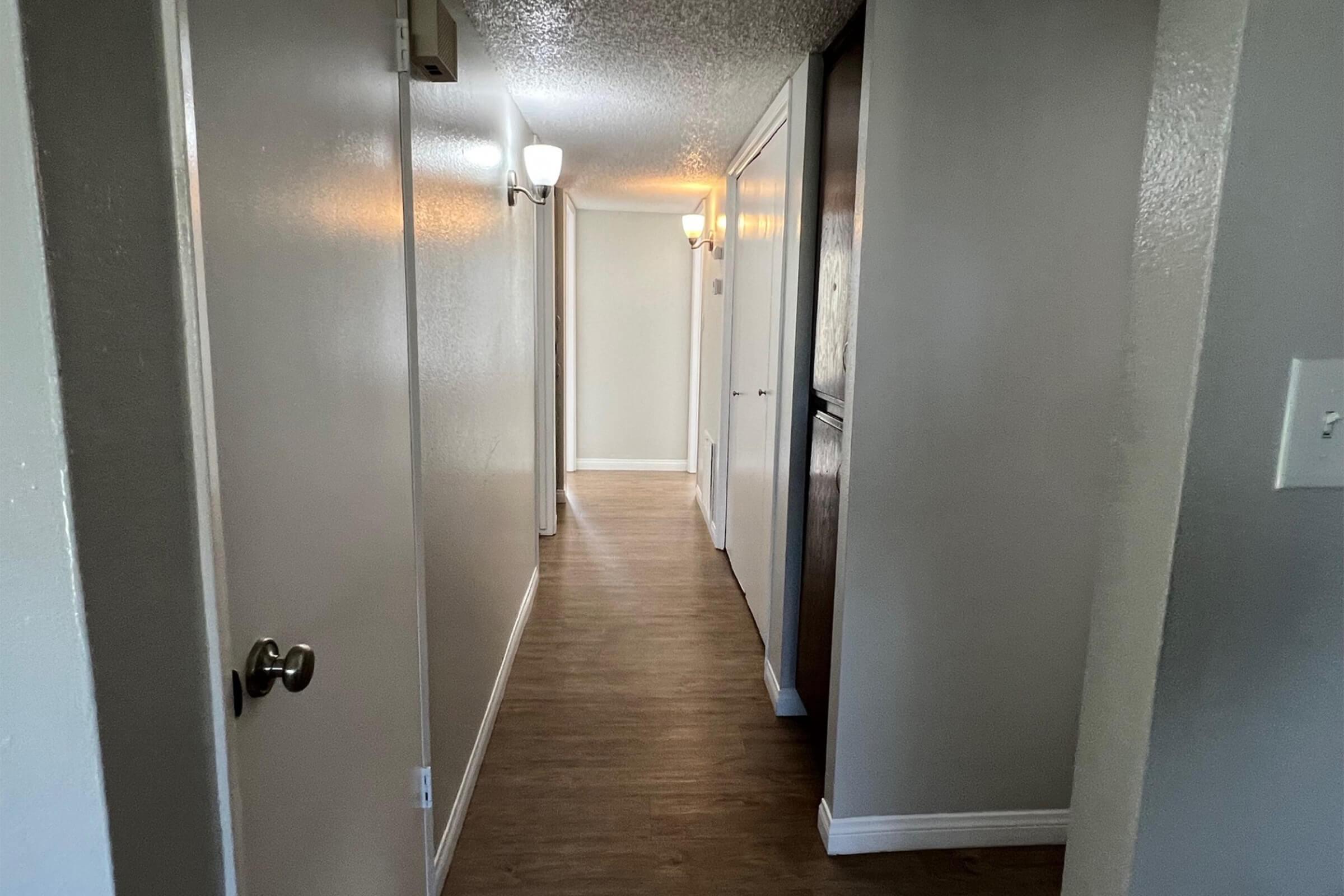 A long hallway with light-colored walls and a wooden floor. There are doors on both sides, with a white closet door visible. Ceiling lights provide illumination, and the space appears clean and well-maintained.