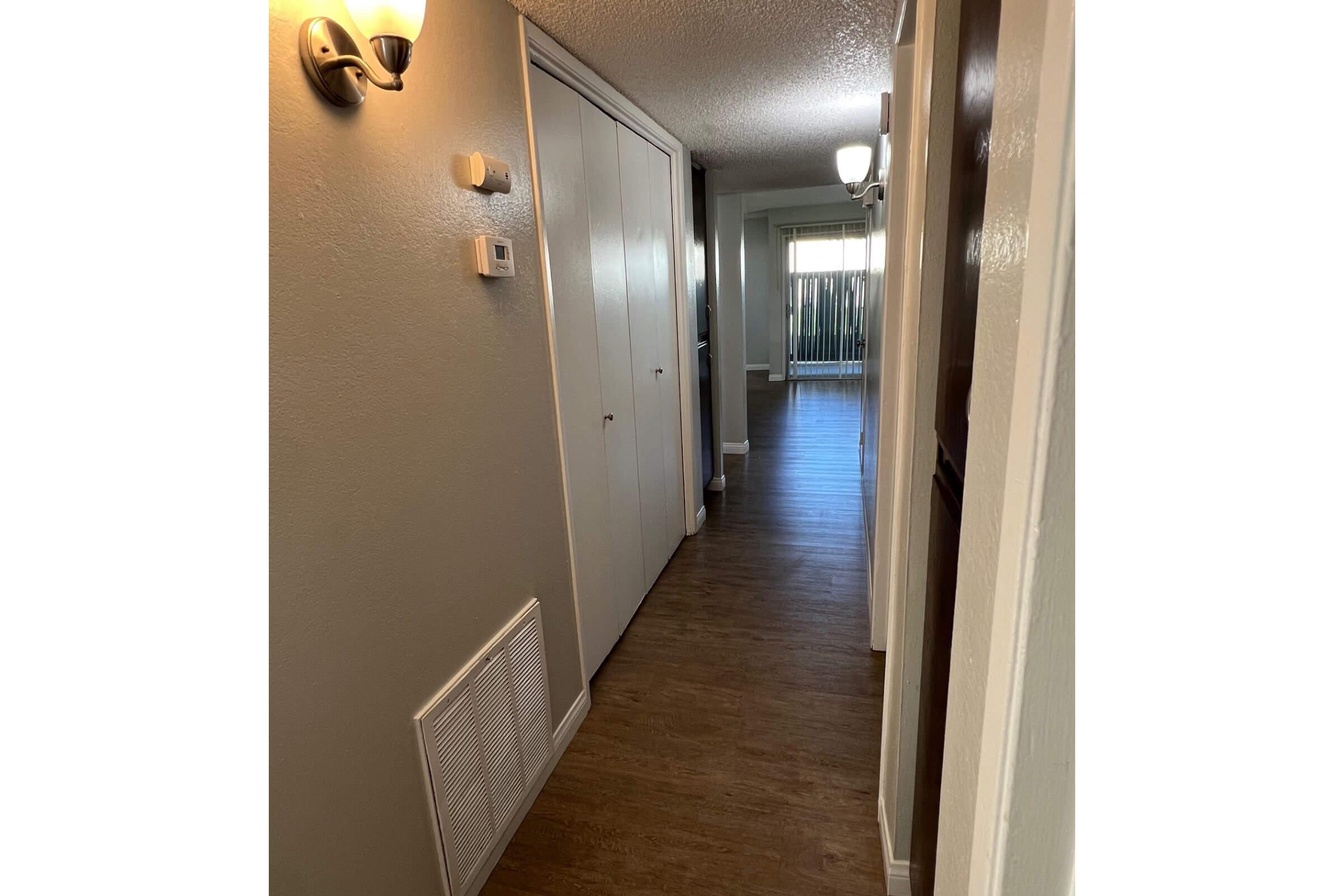 A narrow hallway with light-colored walls and wood flooring. On the left, there is a light fixture and a closet door. The hallway leads to a bright living area visible at the end, with sliding glass doors and additional light fixtures along the ceiling.